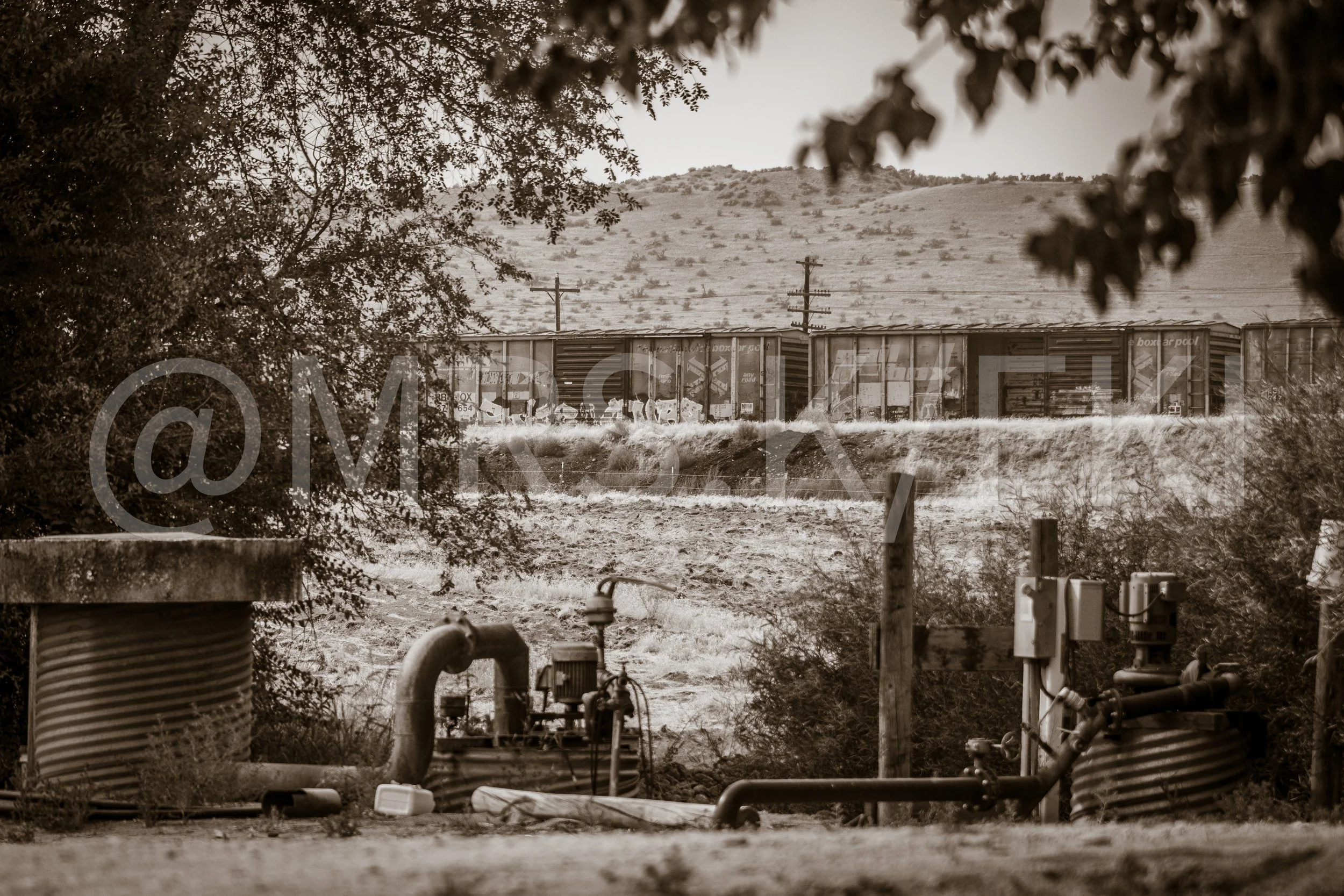 A breathtaking original 1 of 10 sepia-toned image showing a landscape with terrain in the background. In the foreground, there are industrial elements such as large metal pipes, utility boxes, and barrels, with trees framing the scene on both sides.