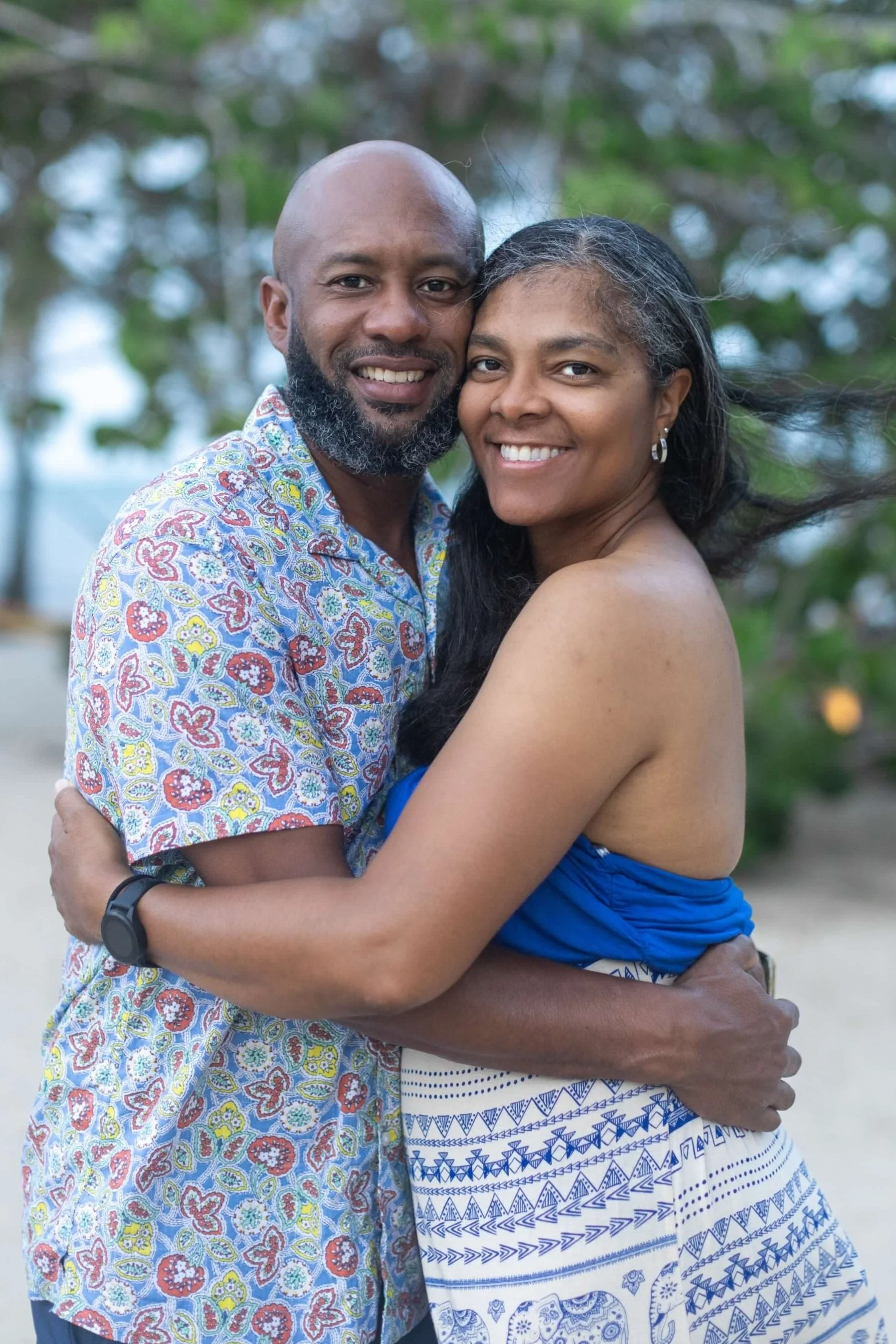 A smiling couple hugging outdoors in front of a tree, with the man wearing a colorful patterned shirt and the woman in a blue top and patterned skirt.
