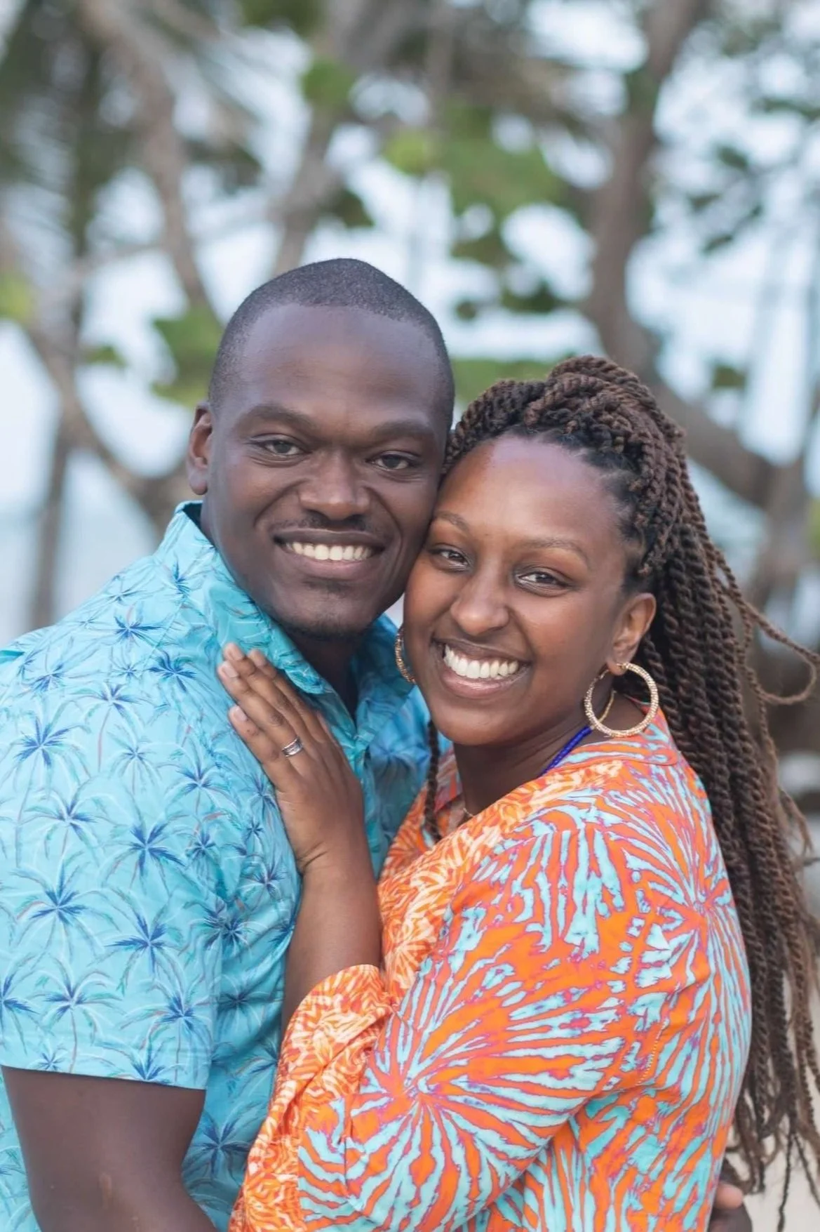 A smiling couple hugging outdoors, with trees in the background. The man is wearing a blue patterned shirt and the woman is wearing an orange and blue patterned dress with large hoop earrings.