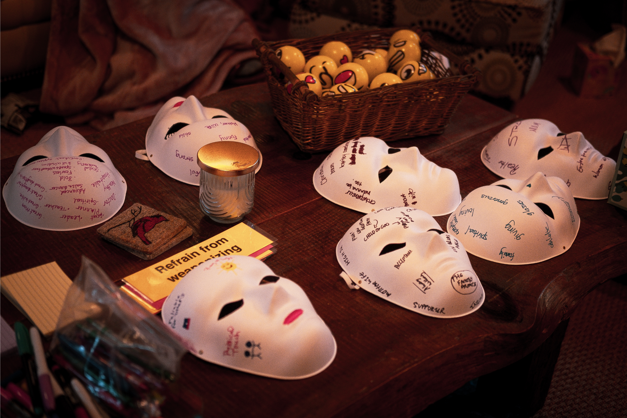 A table with several white masks with handwritten messages on them, a small candle, a basket filled with emoji faces, and some reading materials.