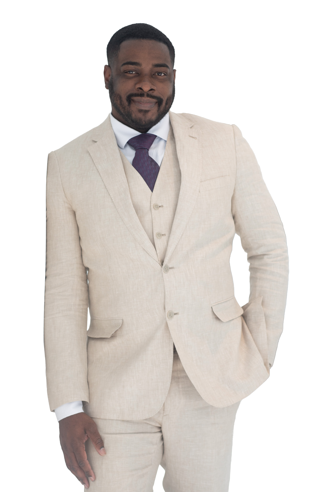 A man in a beige suit with a purple tie, white shirt, and beige blazer, standing with one hand relaxed and the other in his pocket, smiling confidently.