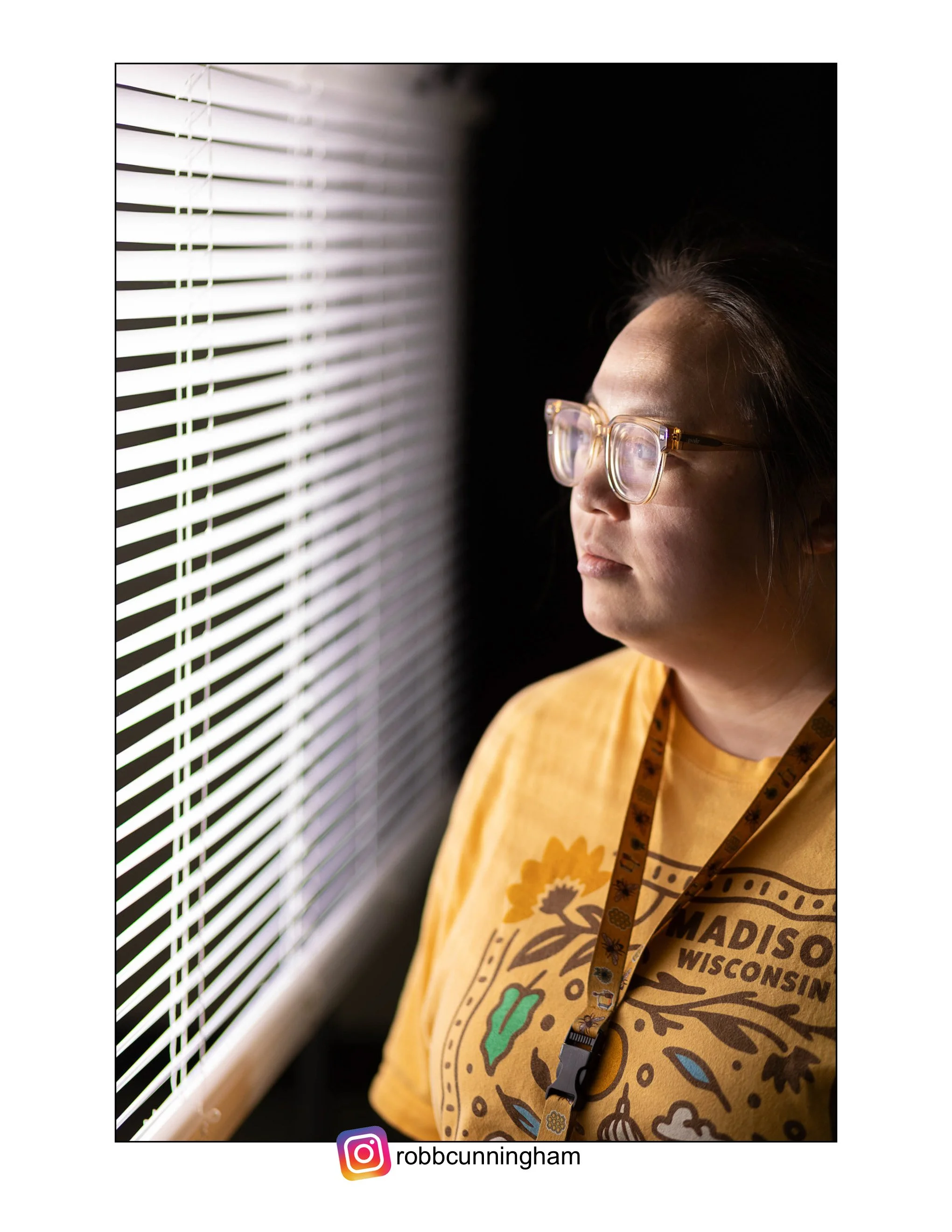 A Hmongwoman wearing glasses and a yellow shirt looks through white window blinds, with a dark background behind her.