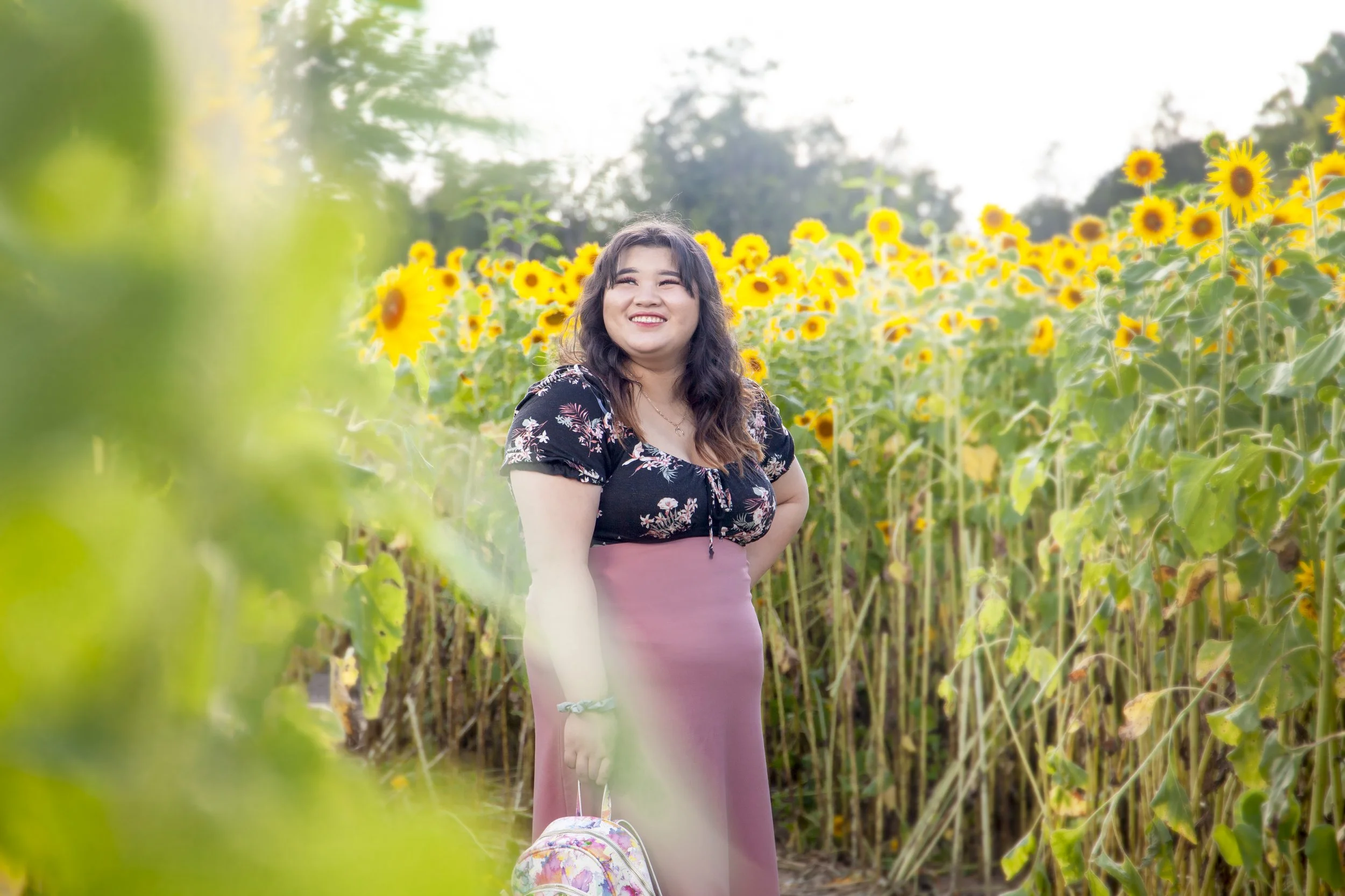 A Hmong woman standing in a sunflower field smiling, holding a small backpack, with green leaves in the foreground and tall sunflowers behind her.