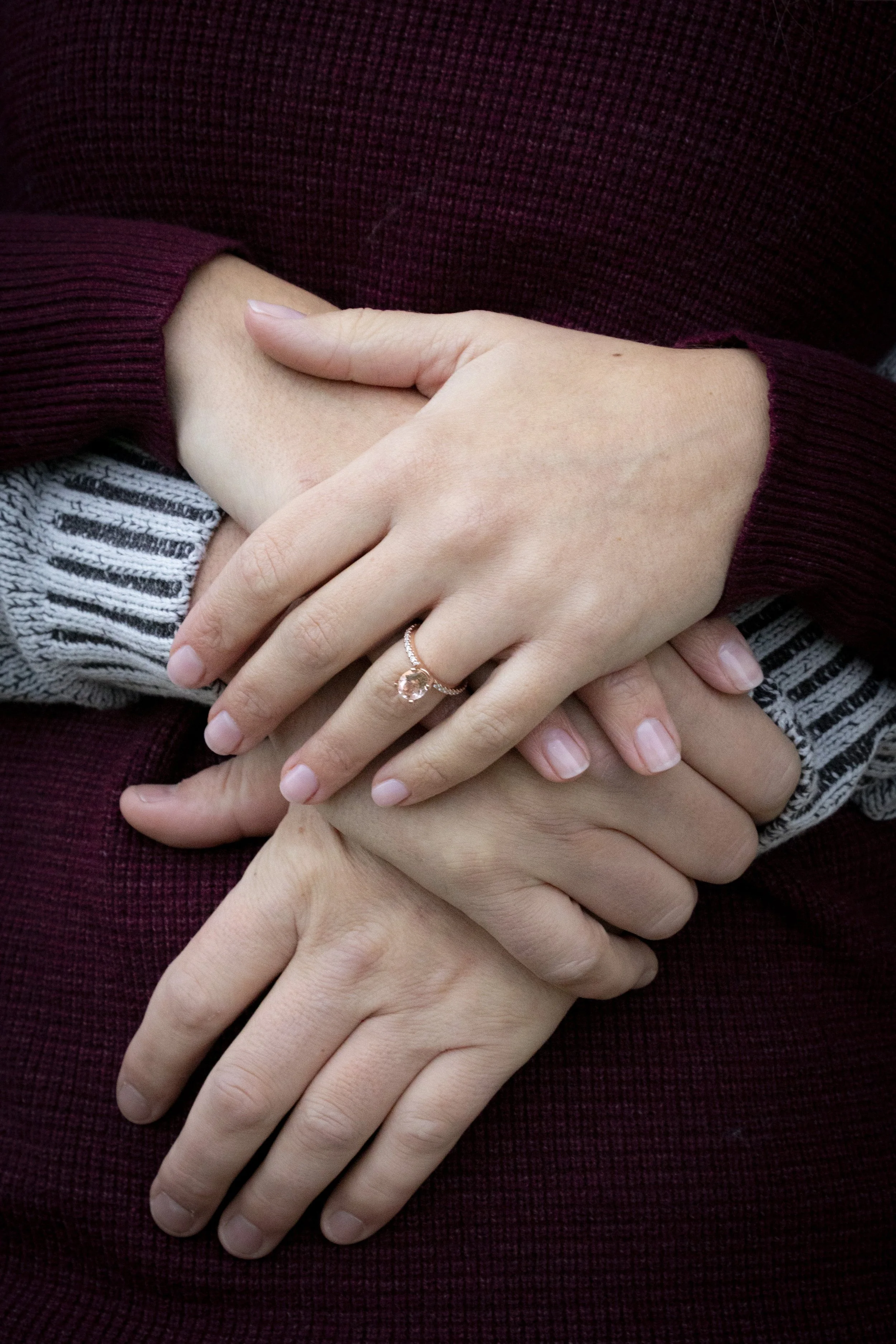 Close-up of two pairs of hands interlocked, one wearing a ring with a pink gemstone, against a background of a maroon sweater and a striped gray shirt.