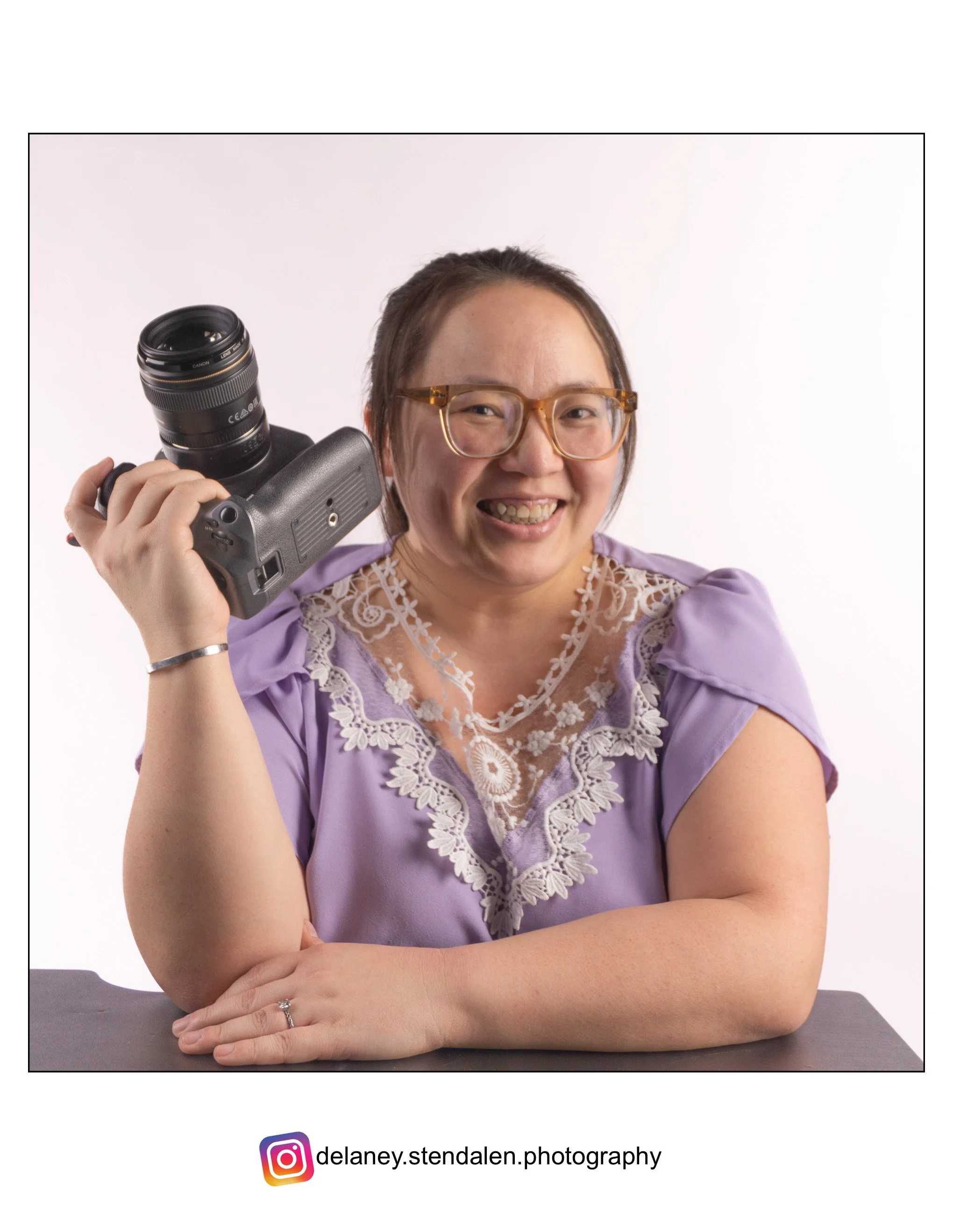 Smiling Hmong woman in glasses holding a camera over her shoulder against a white background.