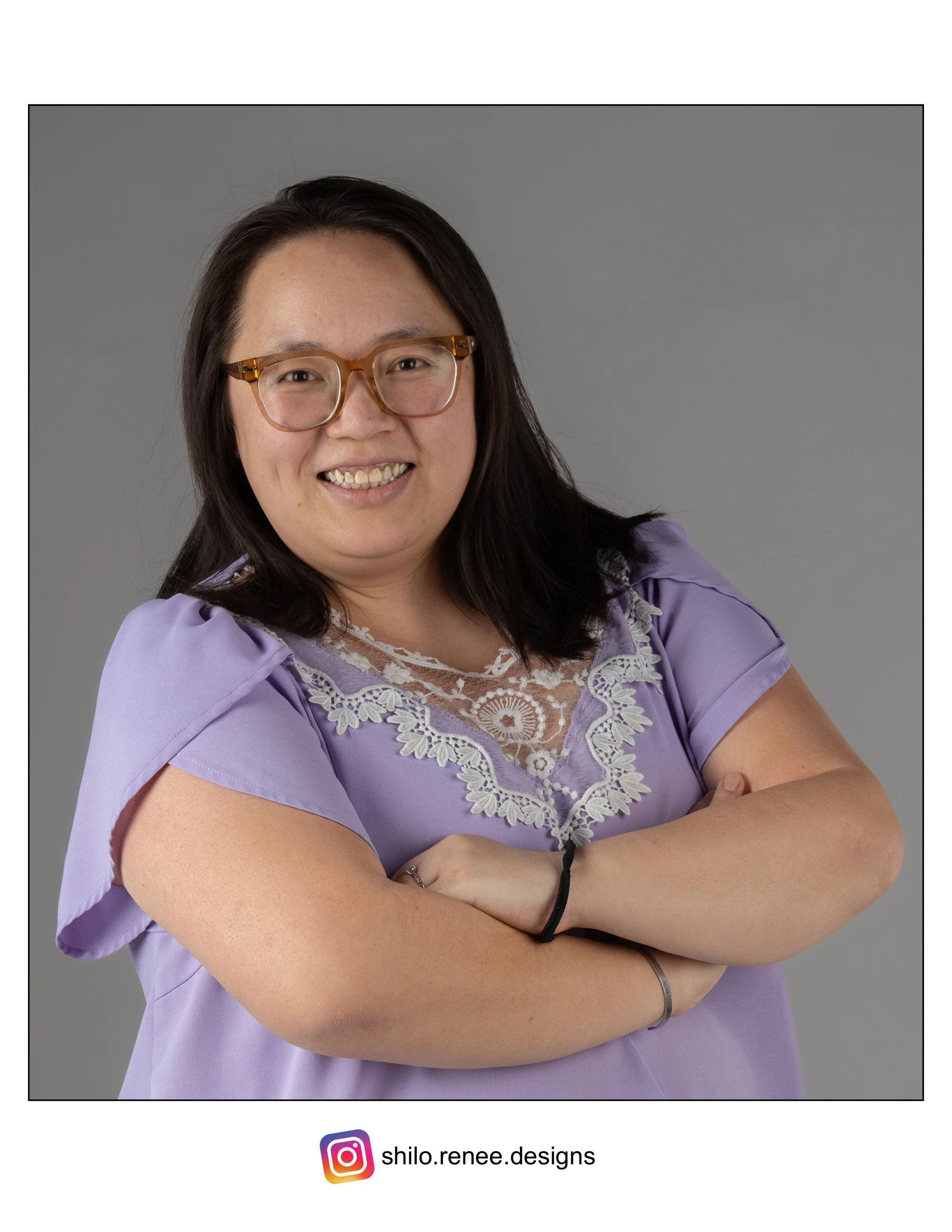 Portrait of a Hmong woman with glasses, wearing a lavender blouse with lace details, smiling with arms crossed against a gray background.
