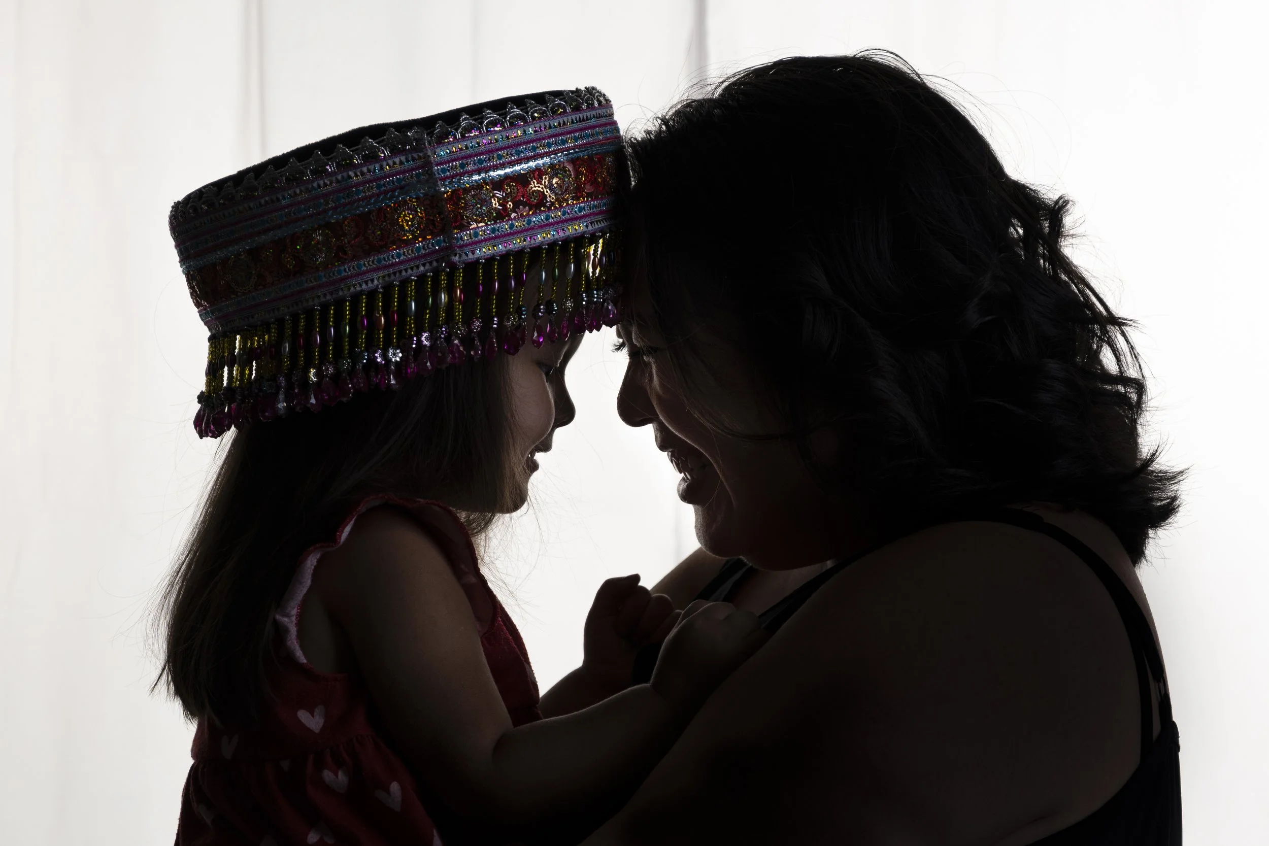 Silhouettes of a mother and a daughter. The girl wears a colorful, beaded Hmong hat and a sleeveless dress with hearts. They are smiling and touching foreheads, showing affection.