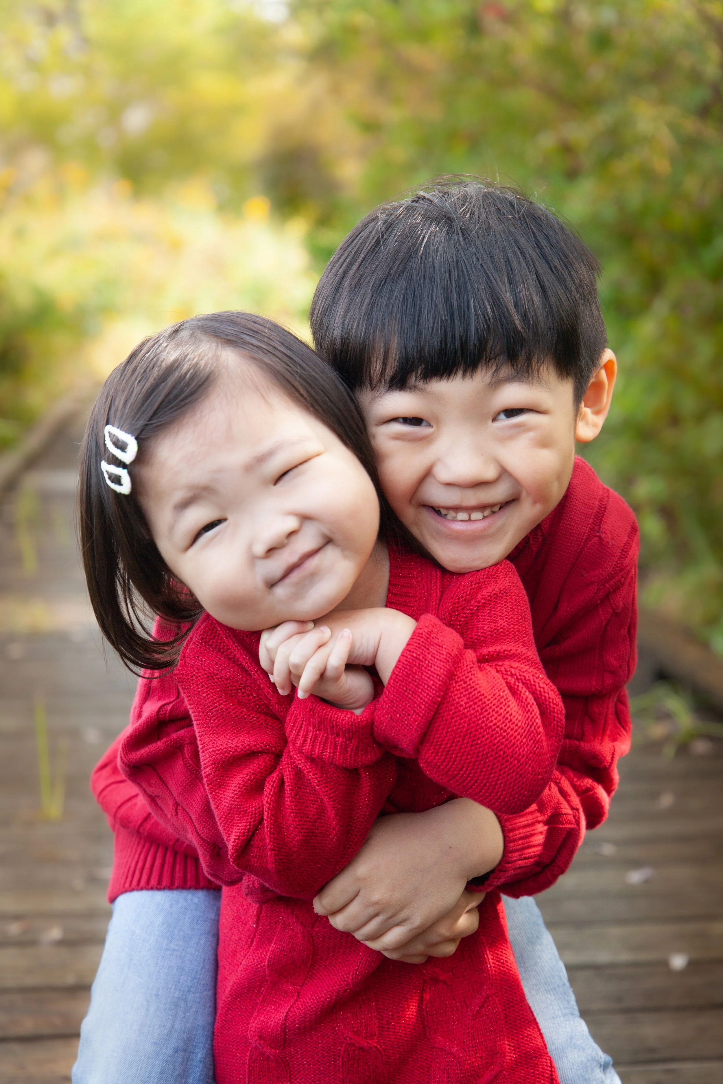 Two Hmong children, a boy and a girl, smiling and hugging each other outdoors on a wooden walkway with green and yellow foliage in the background. Both are wearing red sweaters.