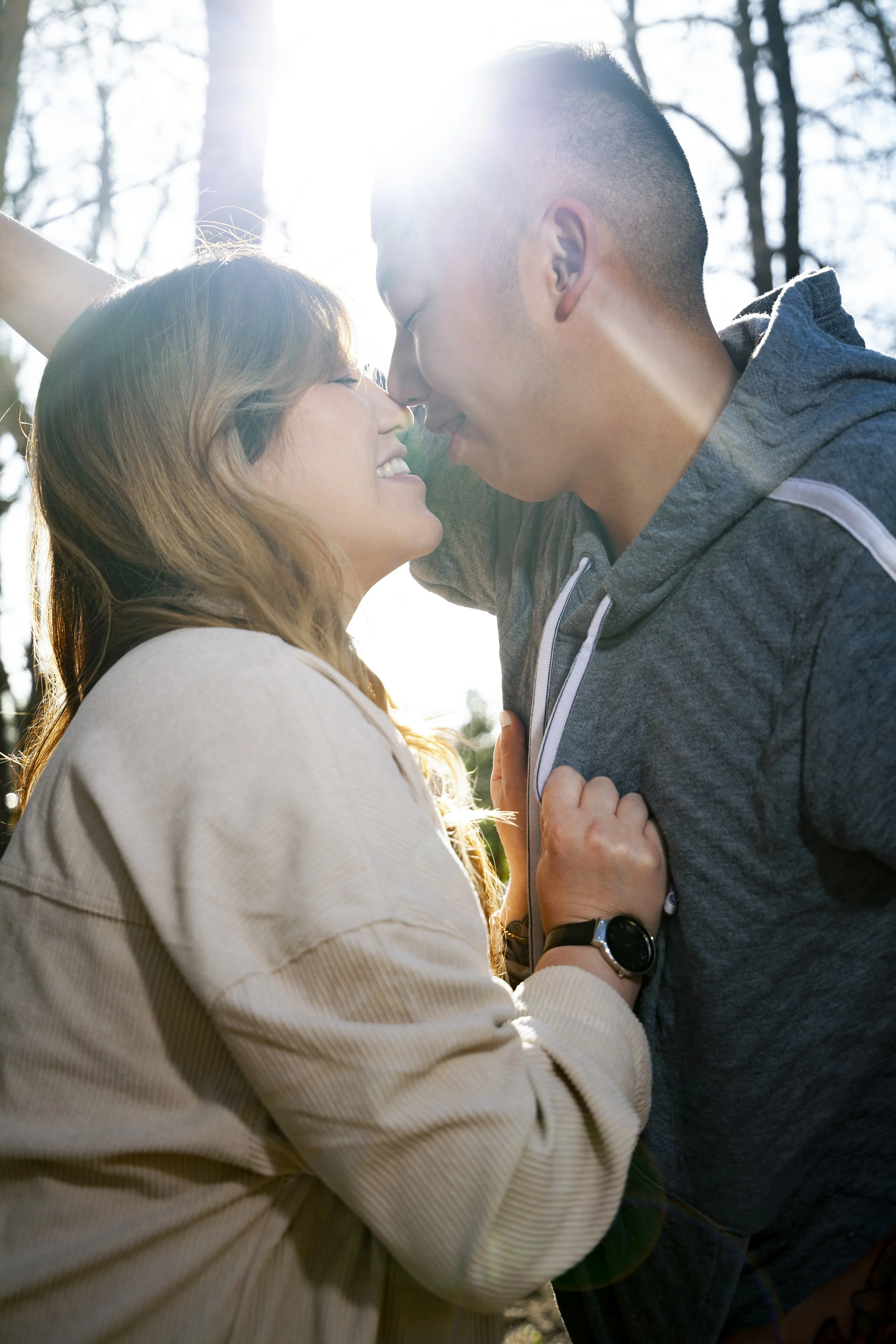 A happy Hmong couple sharing a close moment outdoors with the sun shining through trees in the background.