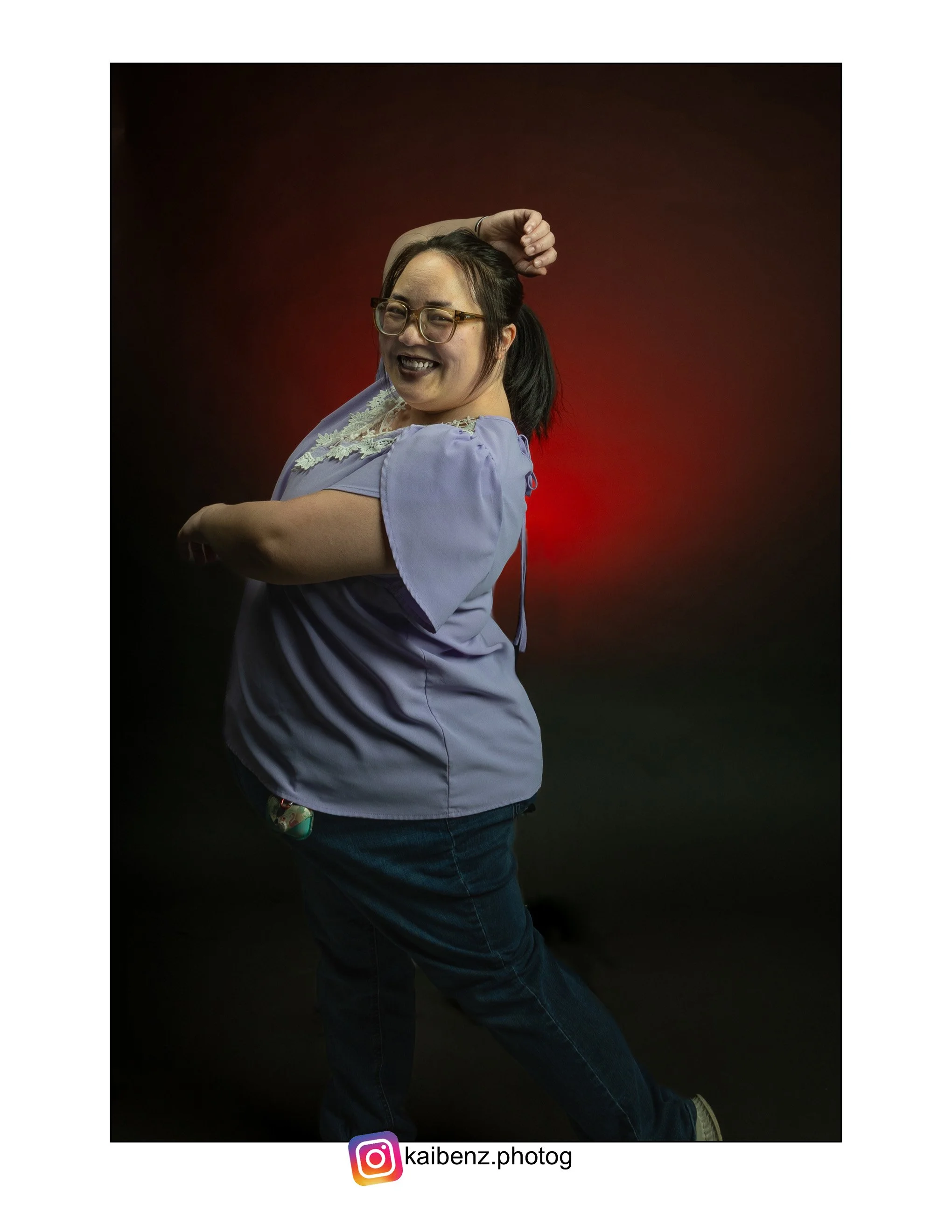 A Hmong woman with glasses, smiling, posing with one arm bent and the other arm raised behind her head, wearing a purple top with lace, standing against a dark background with red lighting.