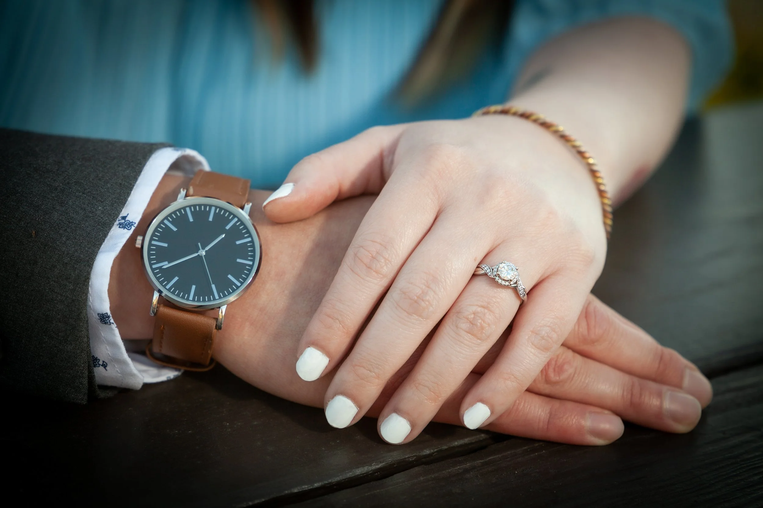 Close-up of a man and woman holding hands on a picnic table, with the woman's hand showing a diamond engagement ring. The man wears a watch with a black dial and a brown leather strap.