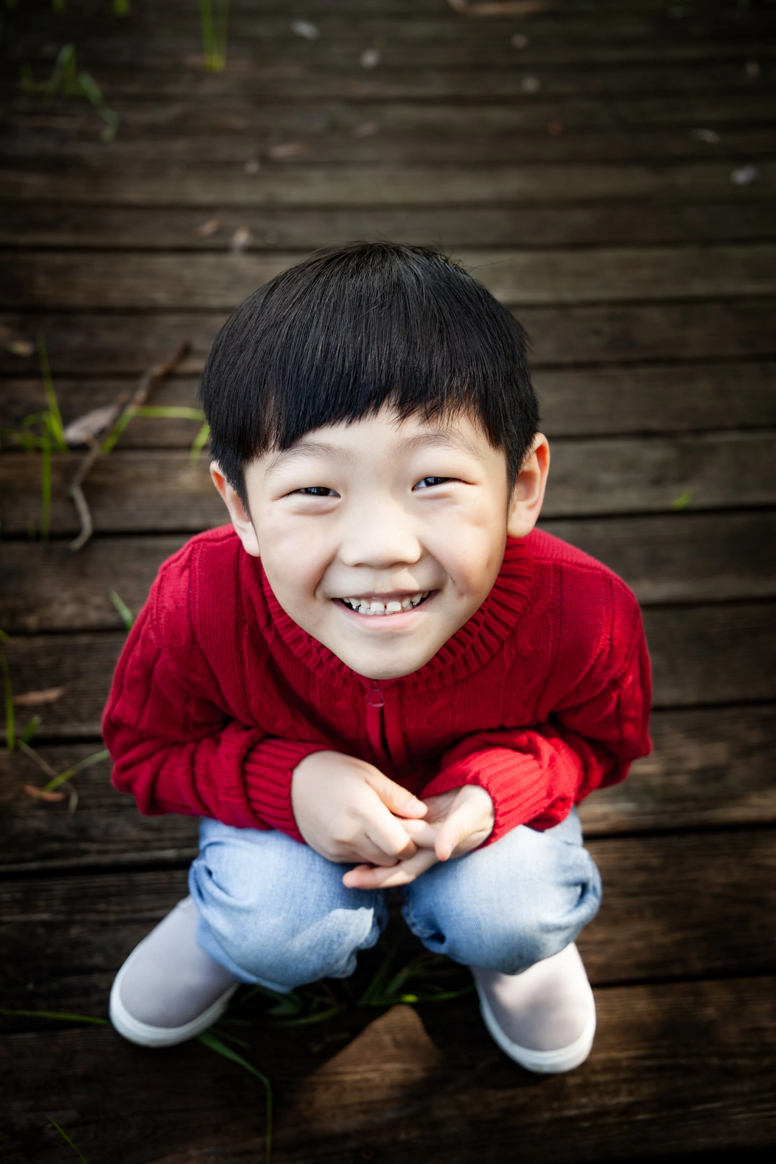 Smiling young Hmong boy with black hair wearing a red sweater and light blue jeans, crouching on a wooden deck outside.
