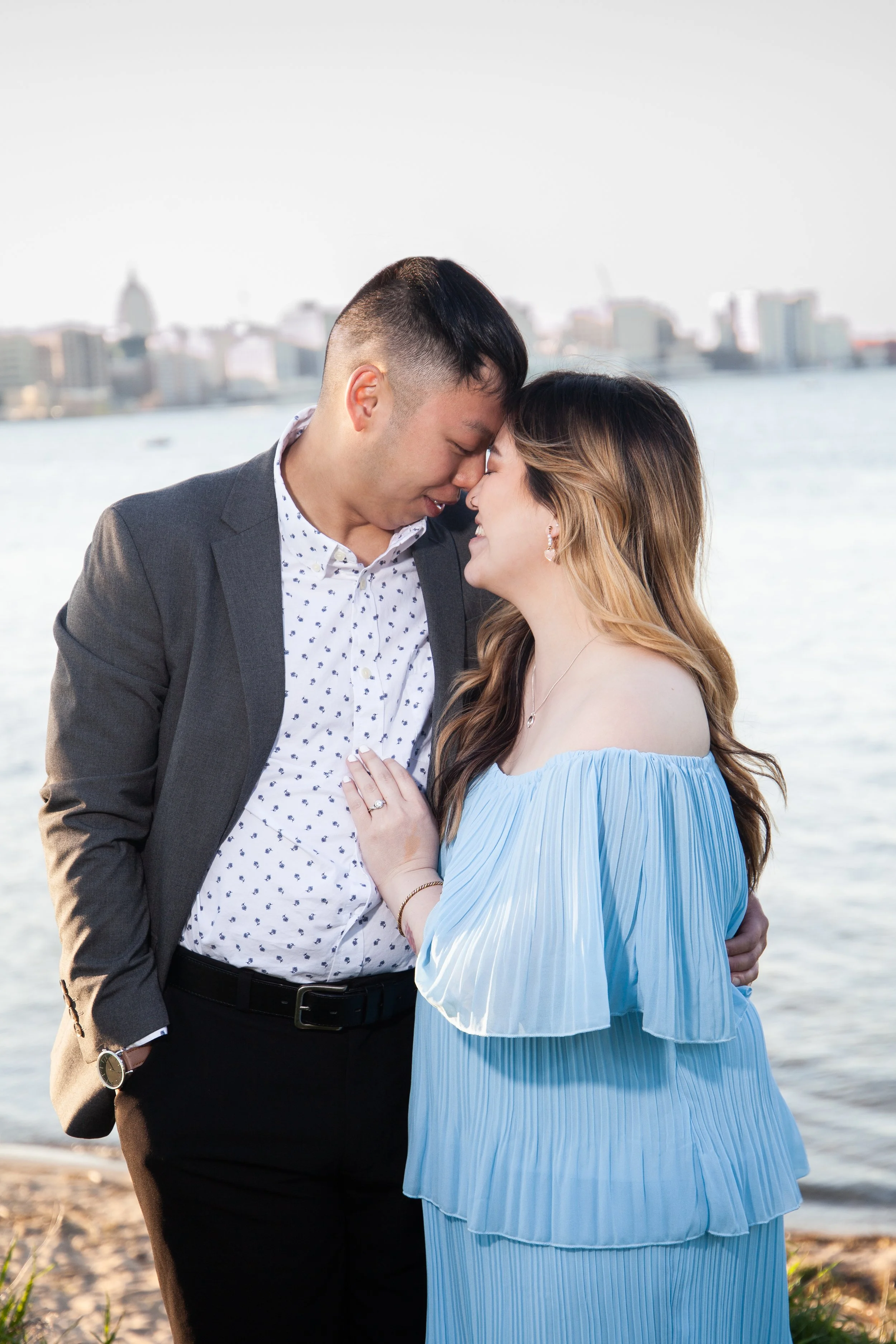 A couple standing close together near a body of water with a city skyline in the background, wearing formal attire and smiling.