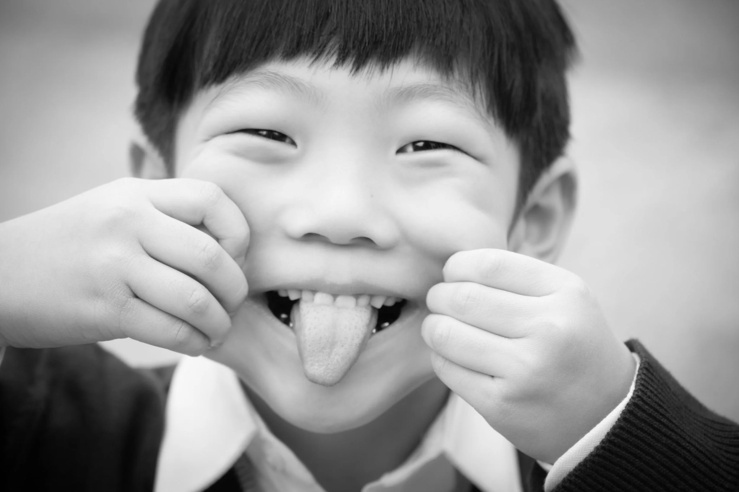 Black and white photo of a Hmong boy playfully sticking out his tongue and pulling his cheeks, smiling with eyes slightly squinted.