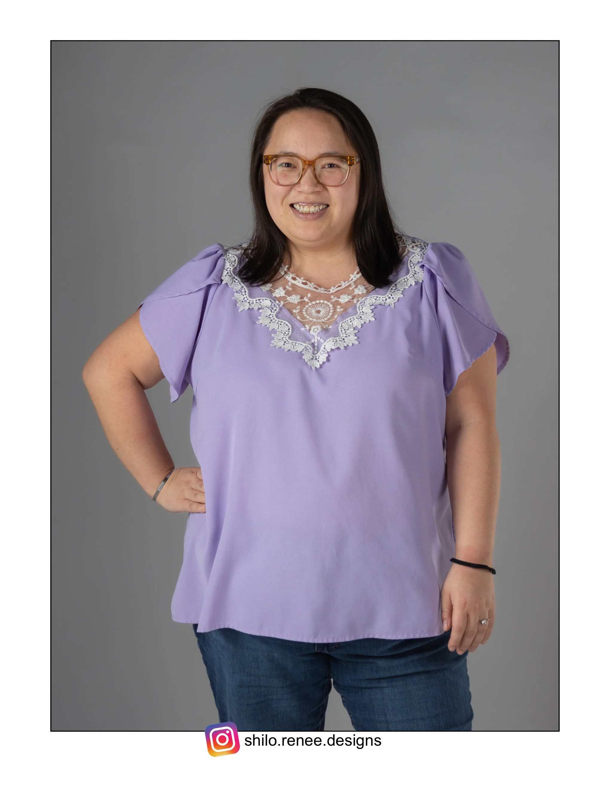 Portrait of a Hmong woman with glasses, smiling, wearing a light purple blouse with lace at the neckline, standing with one hand on her hip against a plain gray background.
