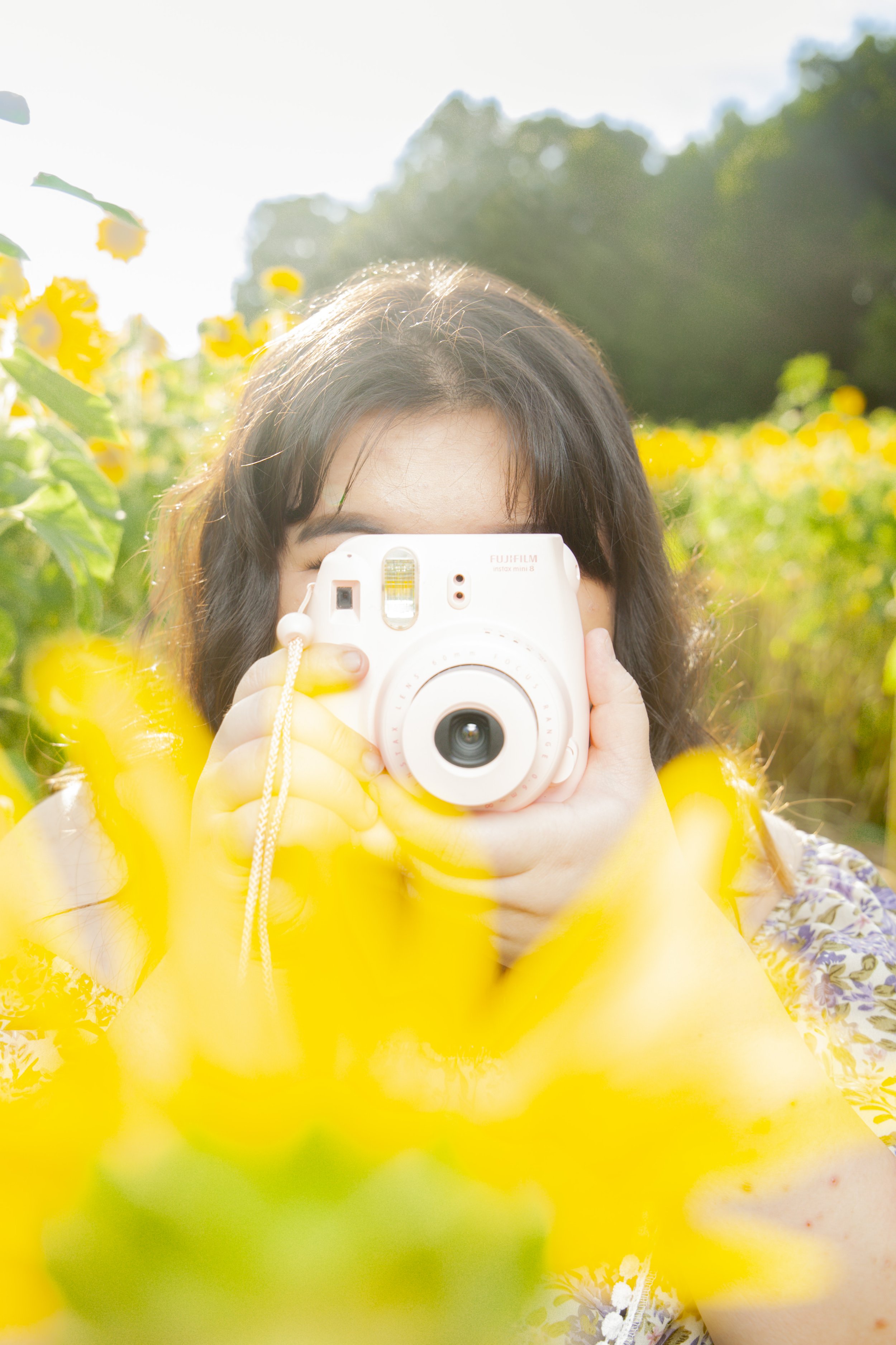 Hmong person taking a photo with a white instant camera in a field of yellow flowers during daytime.