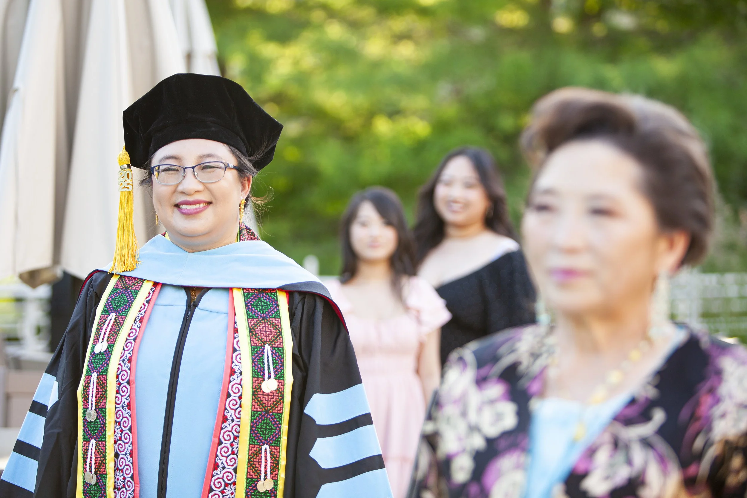 Graduate Hmong woman in cap and gown smiling at outdoor celebration, with two generations of women surrounding her: her daughters in the background and her mother in the foreground.