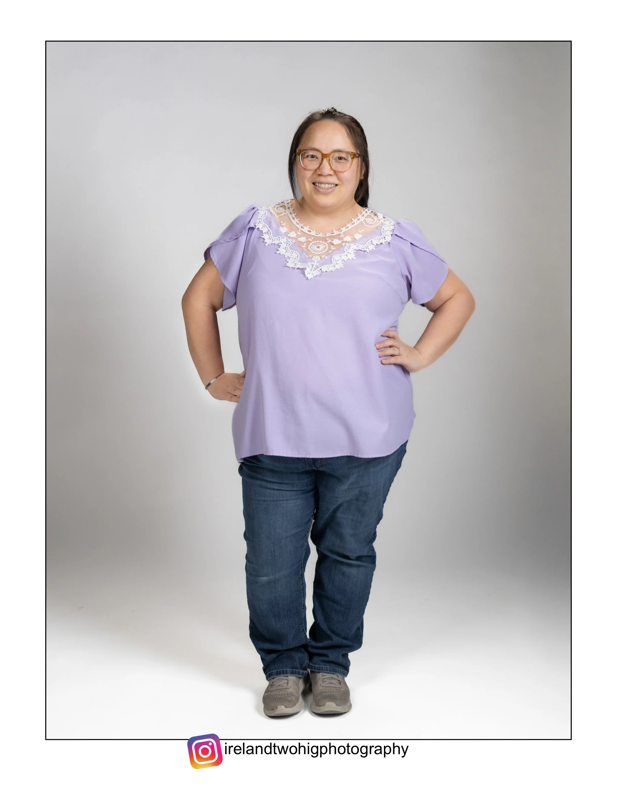 A Hmong woman wearing glasses, a purple top with lace details, jeans, and sneakers, standing with hands on hips and smiling against a plain gray background.
