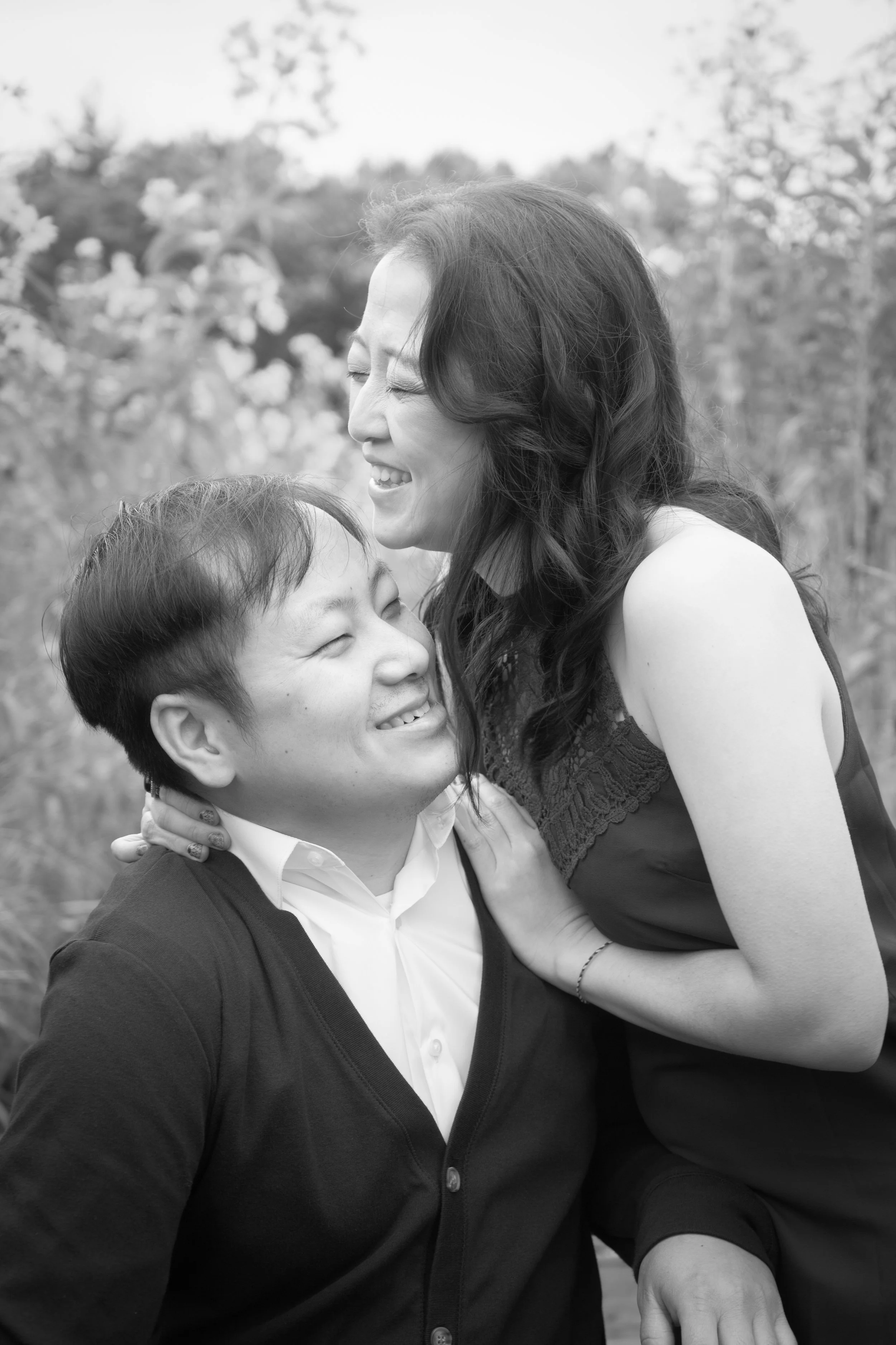 A Hmong woman and a man are sharing a joyful moment outdoors, smiling and embracing with a blurred natural background.