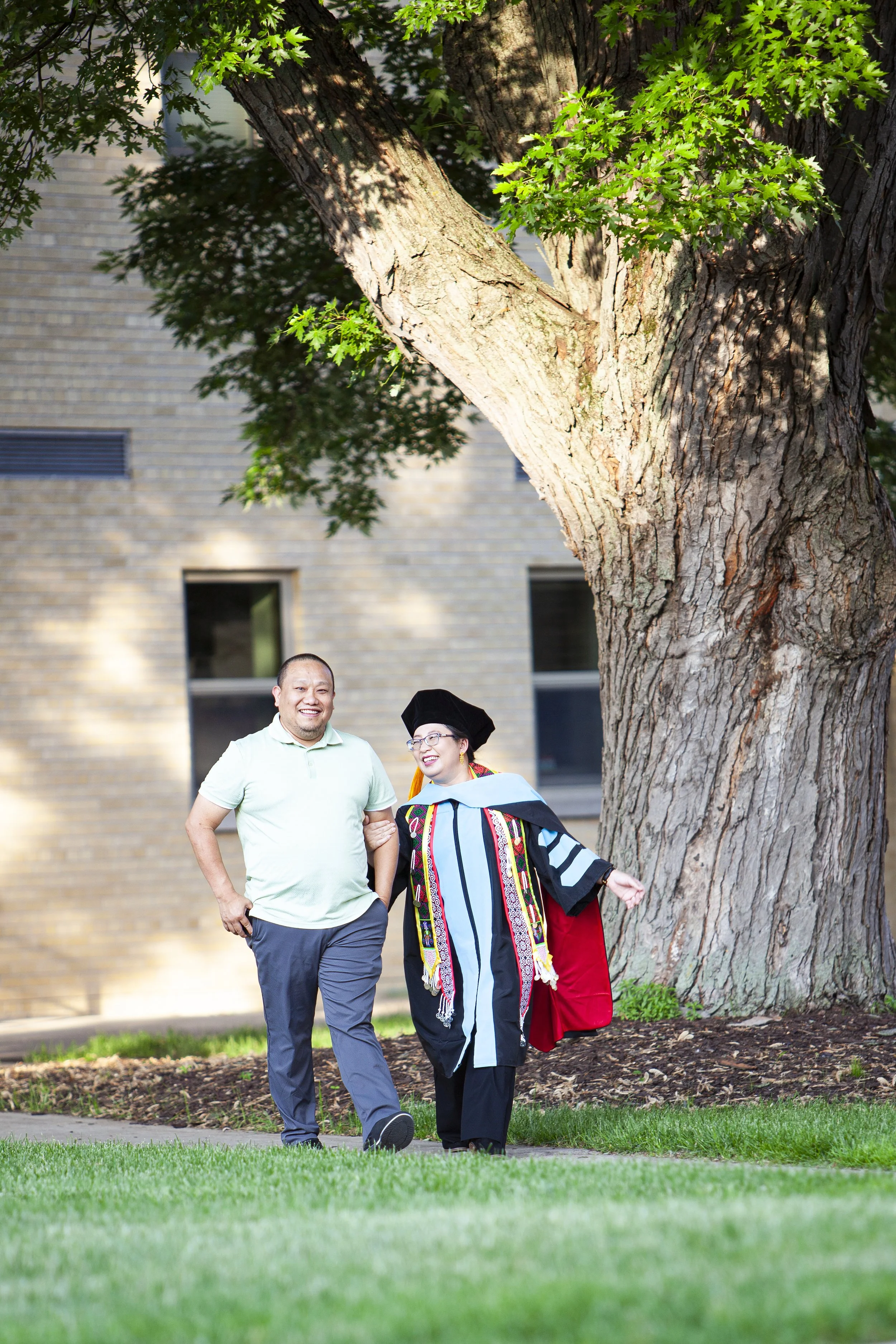 A Hmong graduate in academic regalia walking outside with her husband, smiling, near a large tree and a brick building.