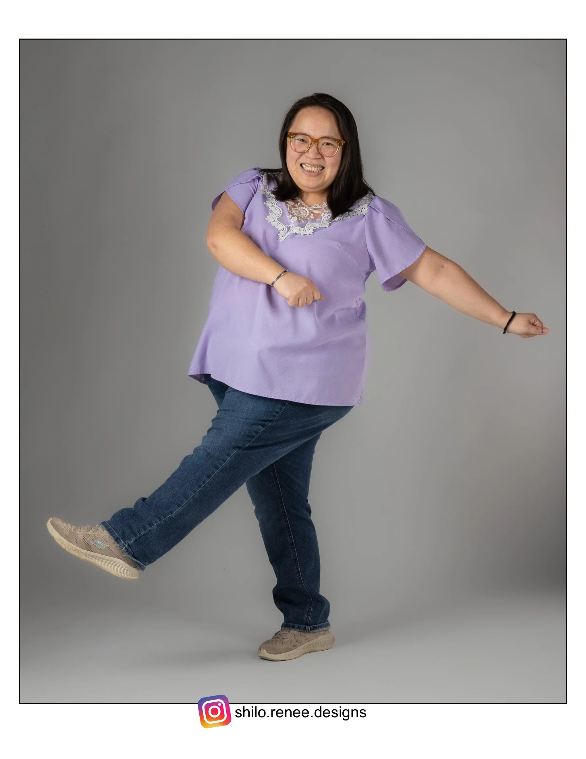A Hmong woman in a light purple top and jeans, smiling and gesturing with her right arm, standing against a gray background.