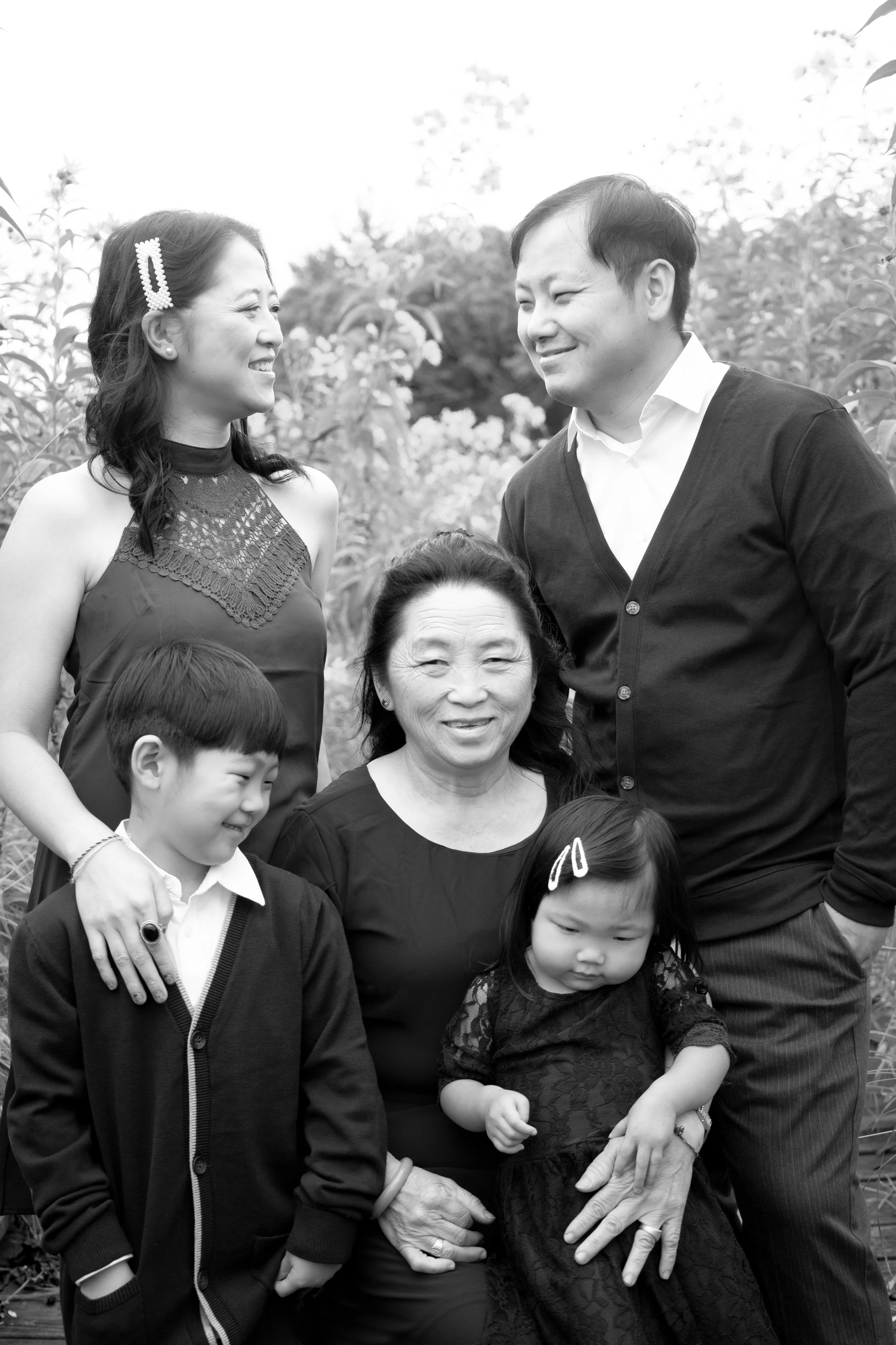 Black and white photo of six Asian family members, three adults and three children, outdoors among trees and plants. The woman in the center sits with a young girl on her lap, while the other woman, man, and children stand around them, smiling and lo