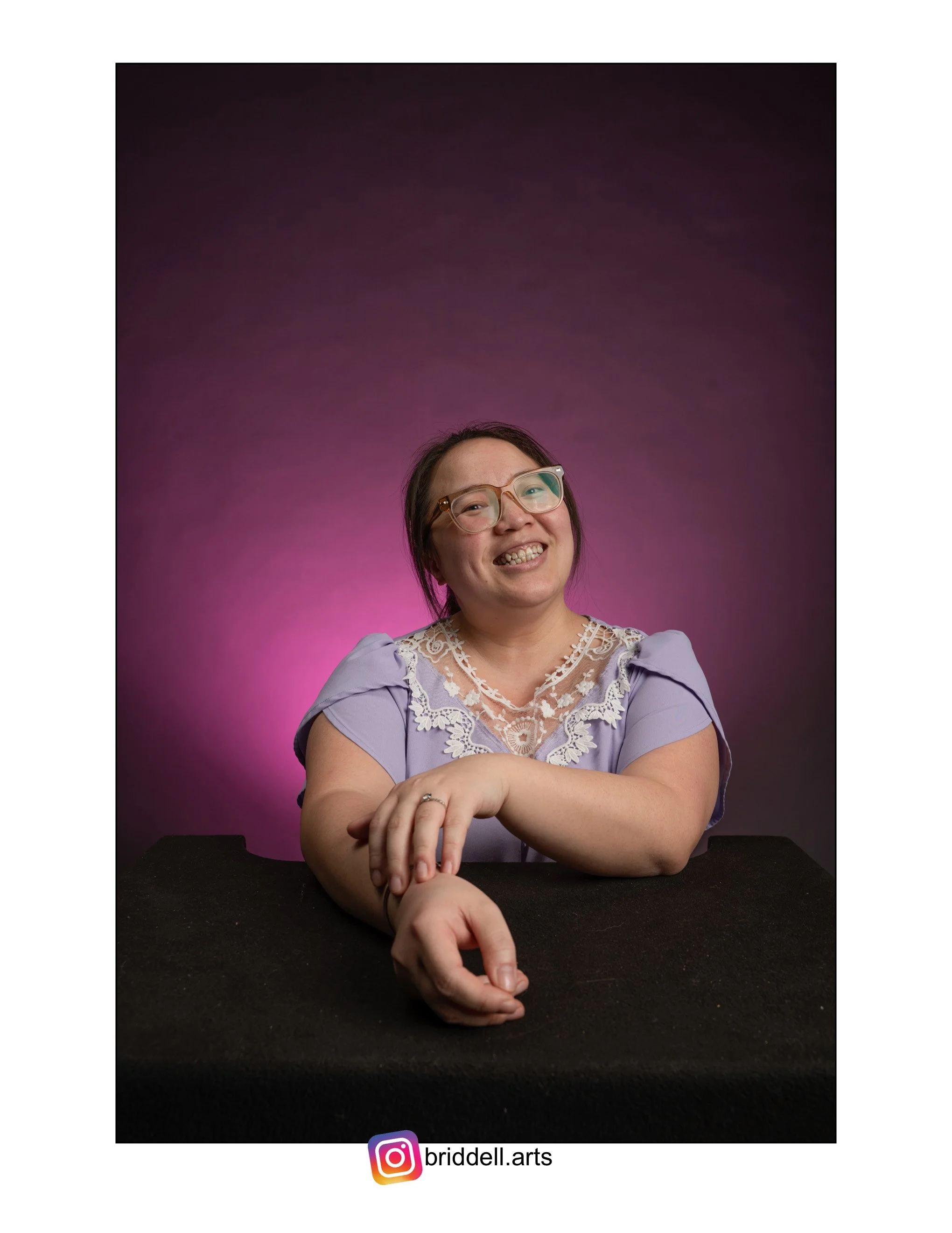 A Hmong woman with glasses smiling, sitting at a black table against a purple background, wearing a lavender dress with lace detail.