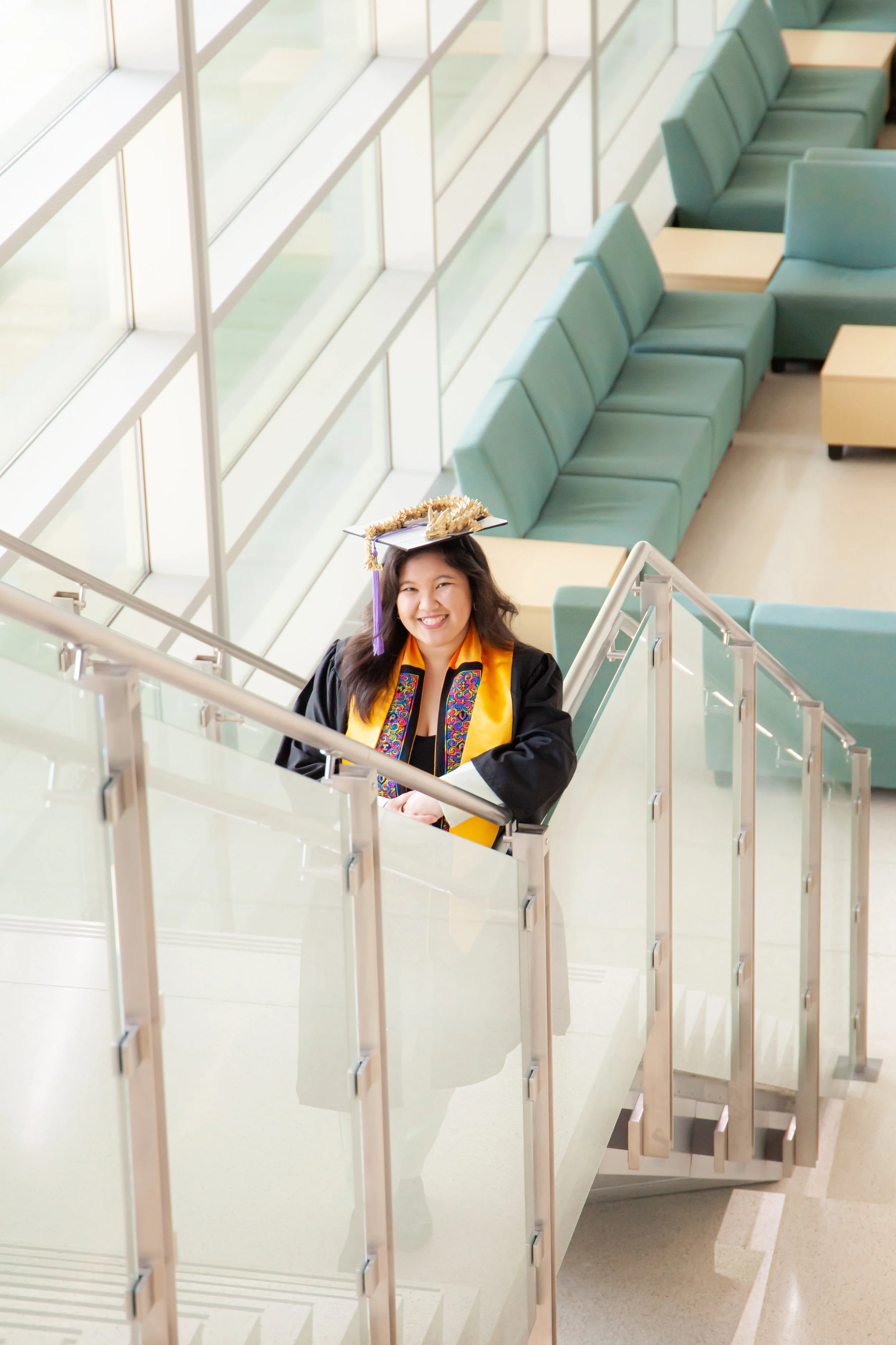 A young Hmong woman in graduation cap and gown with honor cords smiling while standing on a staircase in a modern indoor space with large windows and teal seating.