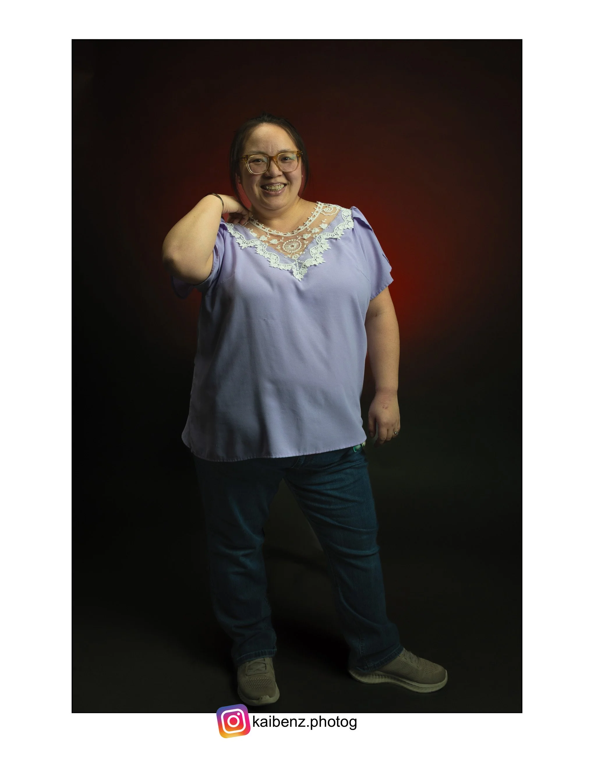A Hmong woman stands against a dark background with a red glow, smiling and posing with one hand behind her head. She is wearing glasses, a purple top with lace details, jeans, and sneakers.
