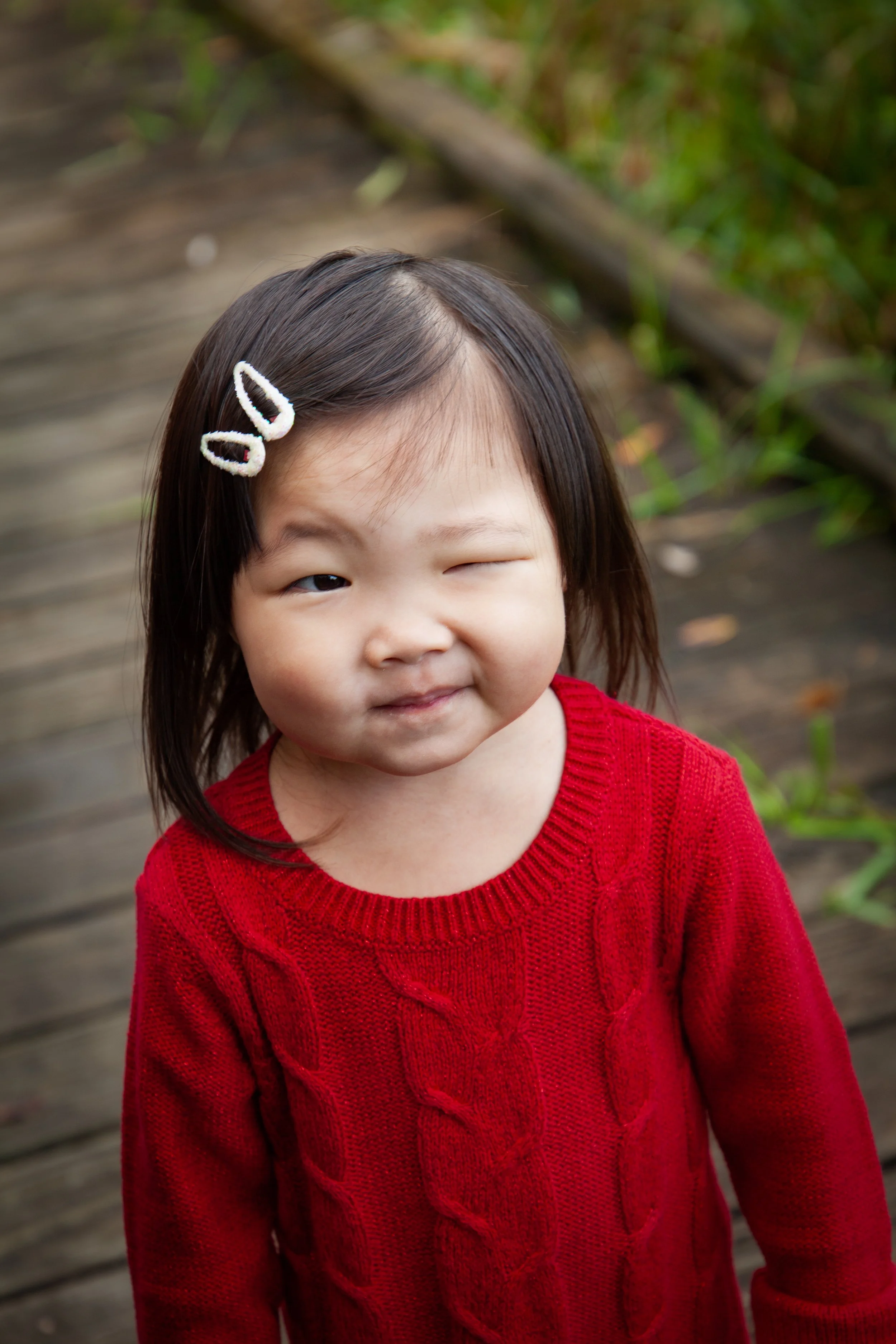 Young Hmong girl with dark hair wearing a red sweater, smiling and winking at the camera, with a wooden walkway and green plants in the background.
