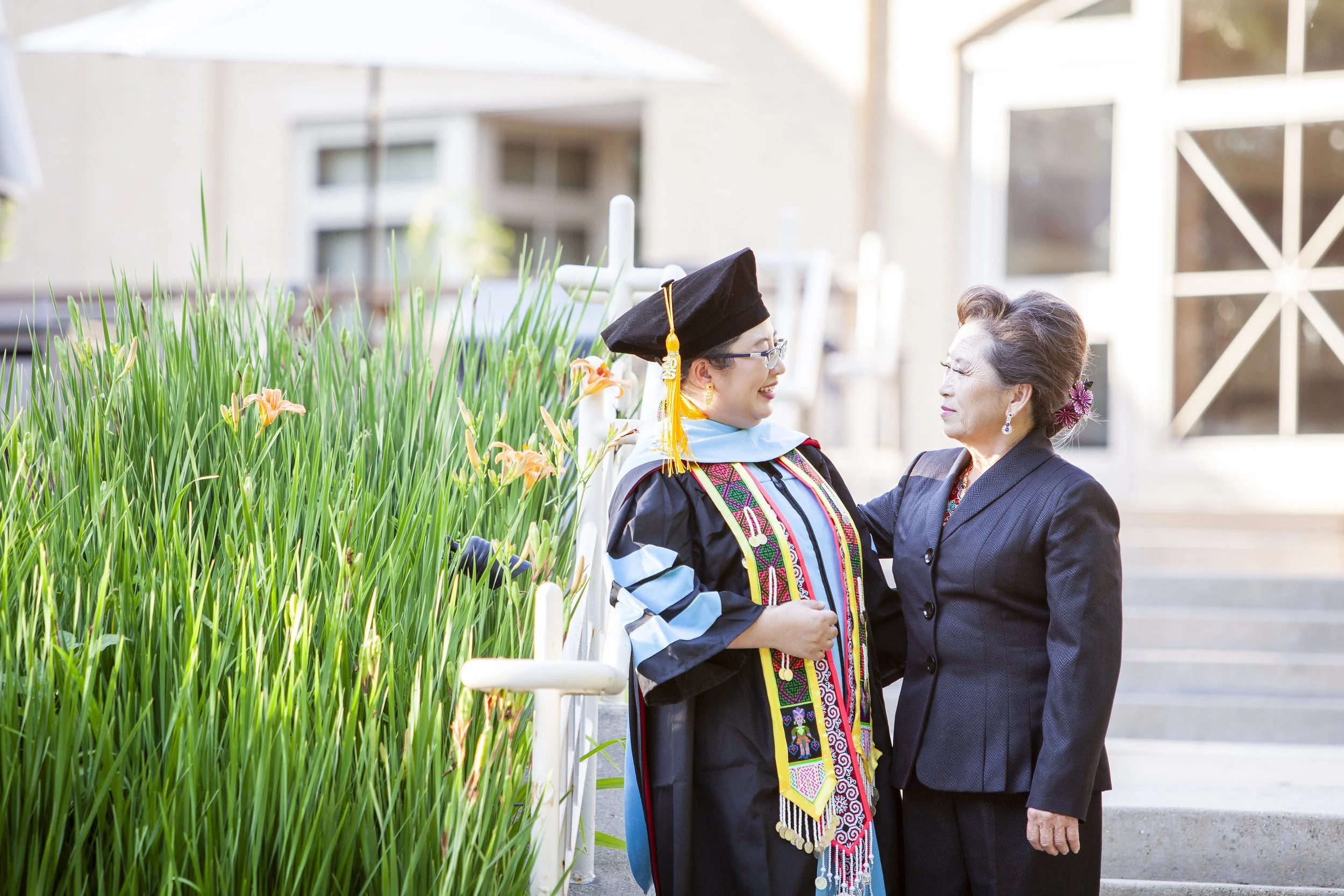 A Hmong woman in a graduation gown and cap standing outdoors on steps, talking to her mother in a dark blazer; both women are smiling and appear to be engaged in a joyful conversation, with green plants and a building in the background.