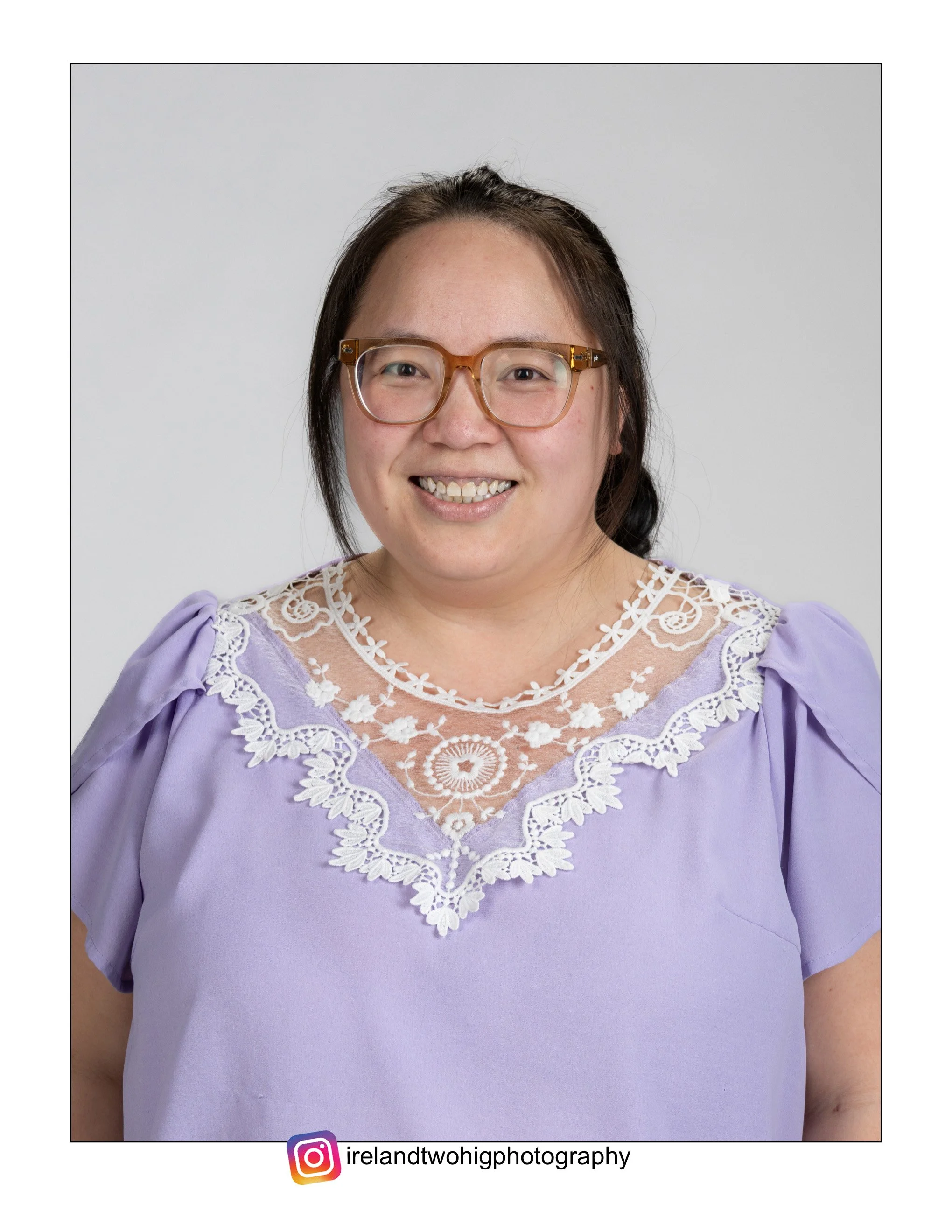 Portrait of a Hmong woman with glasses smiling, wearing a lavender dress with lace detail, against a light gray background.