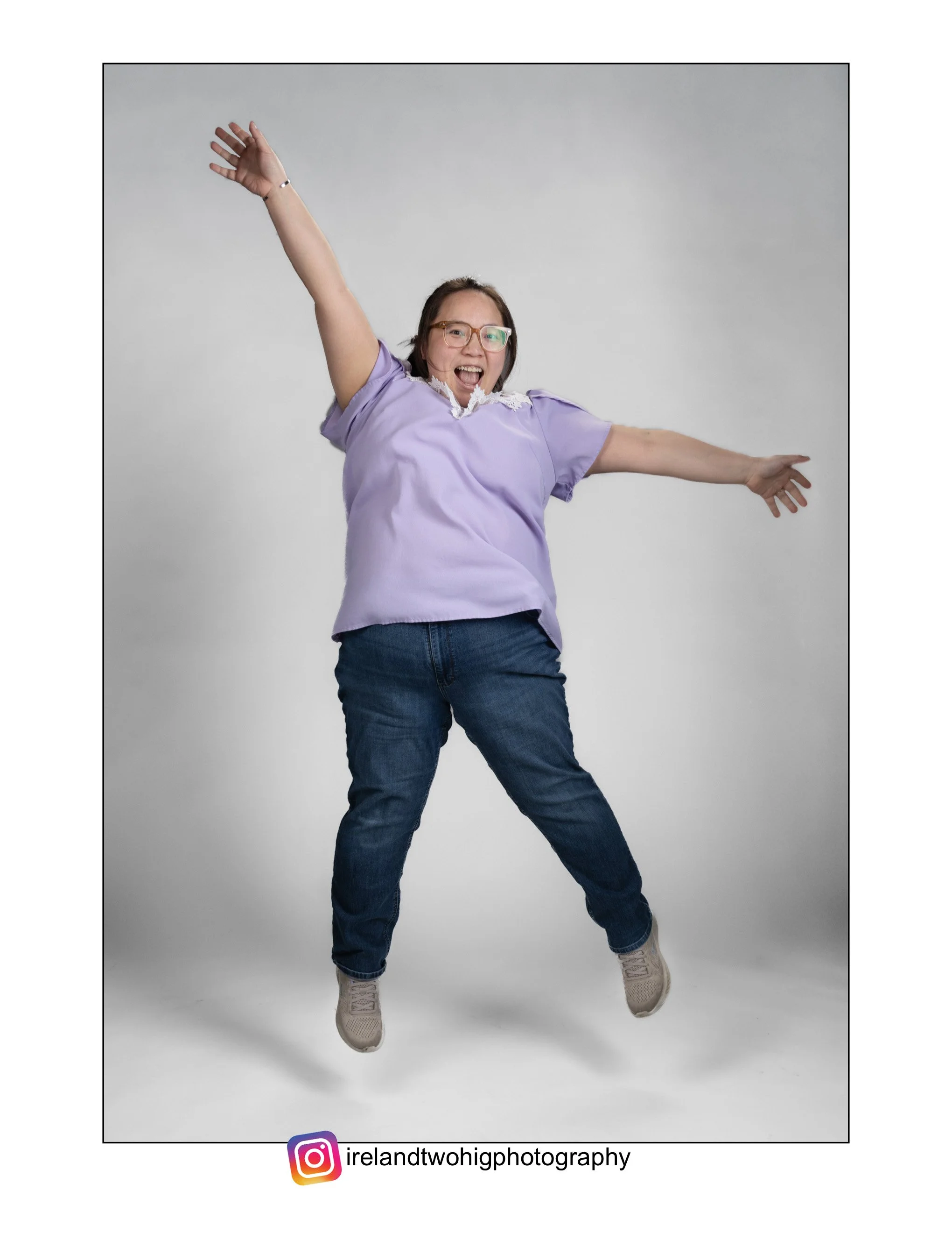 A Hmong woman with glasses, wearing a lavender blouse and jeans, is jumping and smiling against a plain background. She has her arms raised in excitement, caught in mid-jump.