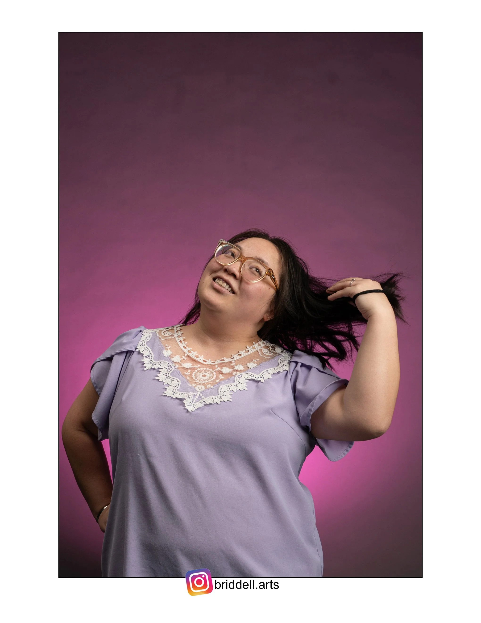 Portrait of a Hmong woman with glasses in a lavender dress with lace details, standing against a purple gradient background, flipping her long dark hair, smiling and posing confidently.