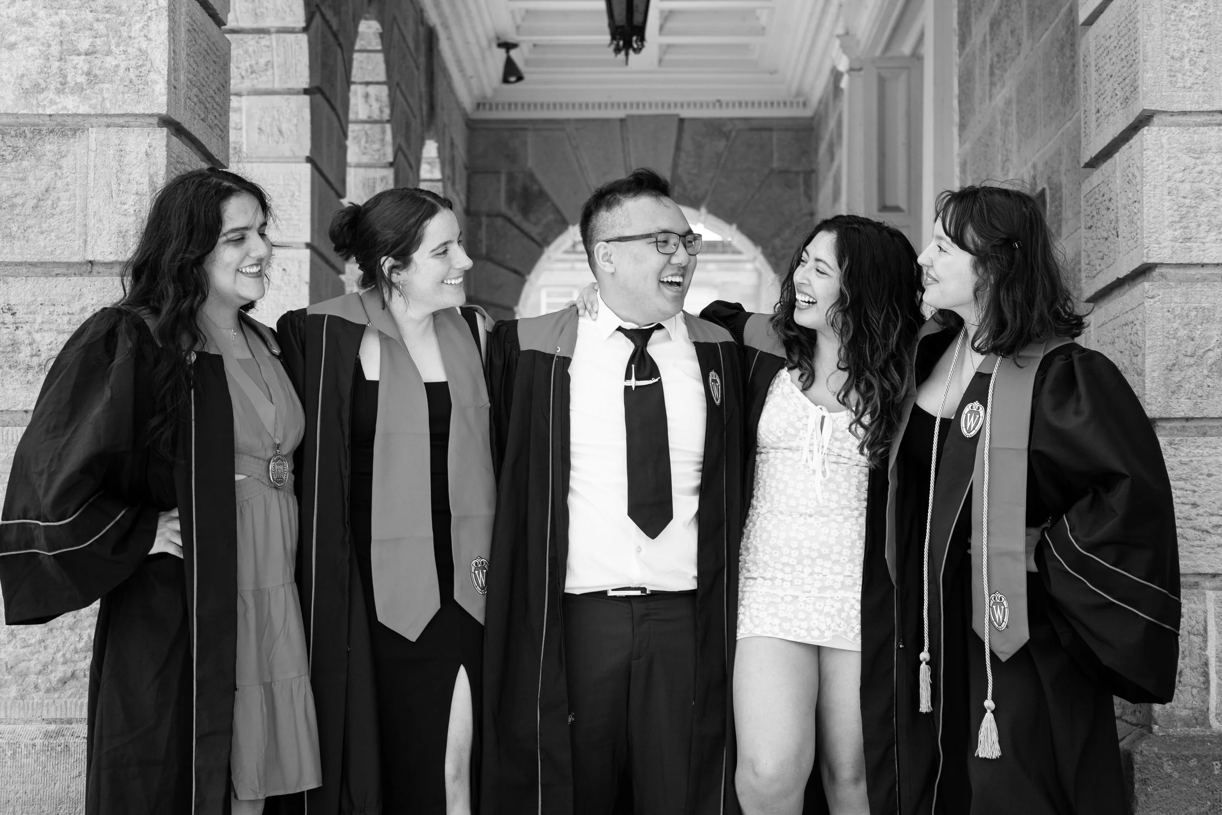 Group of five people of different ethnicities celebrating graduation, wearing caps and gowns, smiling and hugging each other in an outdoor corridor.