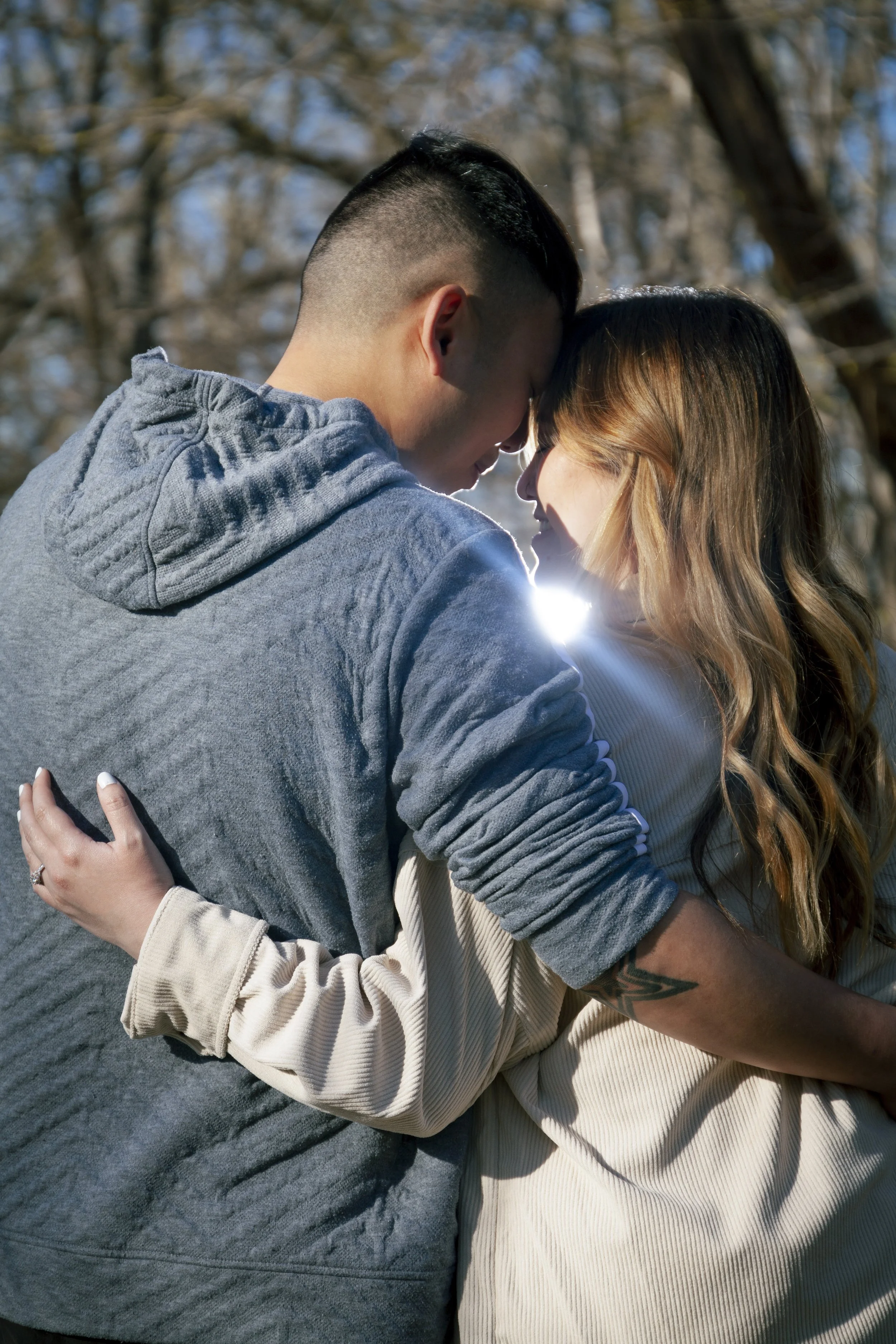 A Hmong couple embracing outdoors, faces close with foreheads touching, sunlight shining behind them, trees in the background.