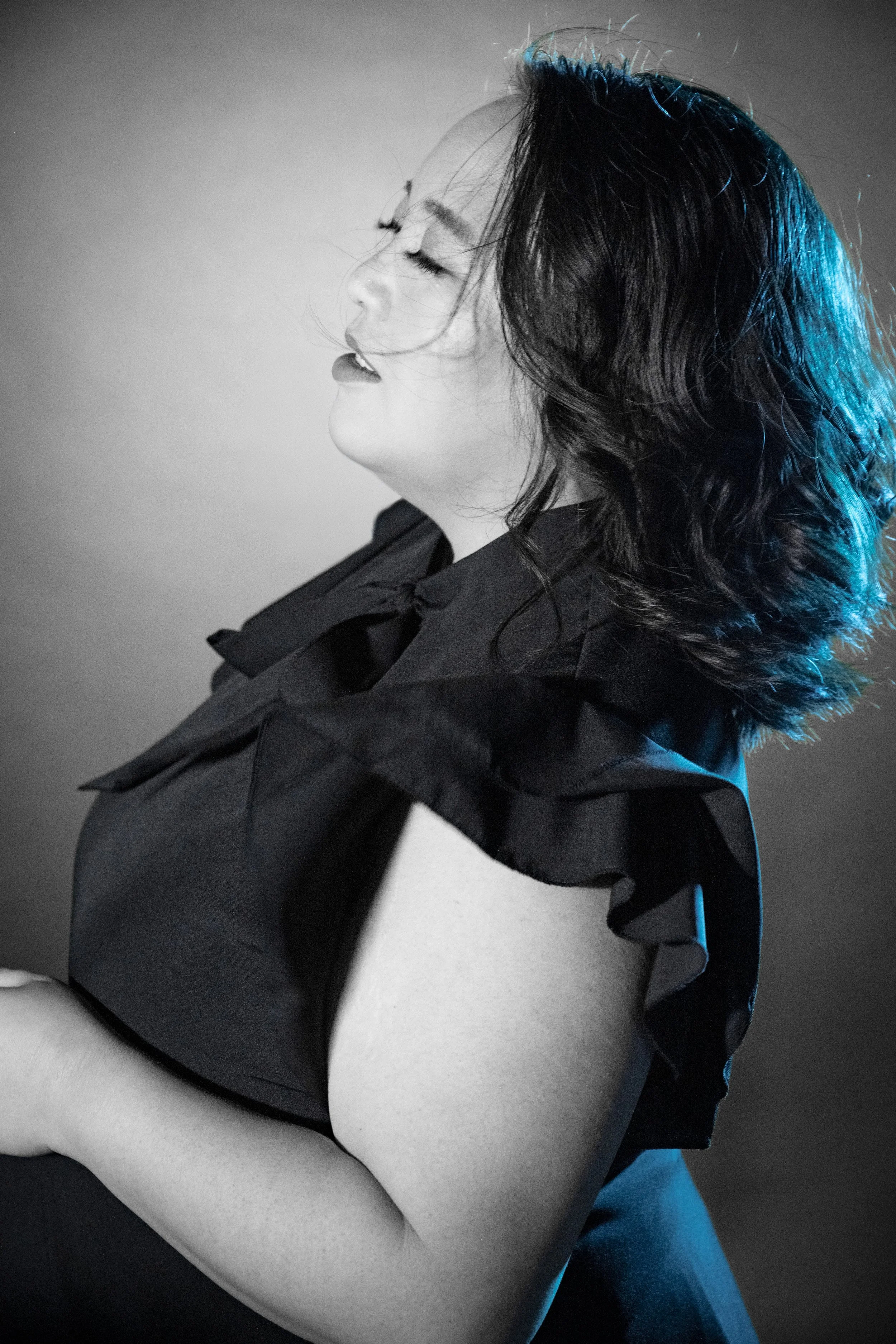 Black and white portrait of a Hmong woman with eyes closed, head tilted back, wearing a sleeveless dress with ruffles on the shoulder, illuminated with soft light and a hint of blue highlight on the hair.