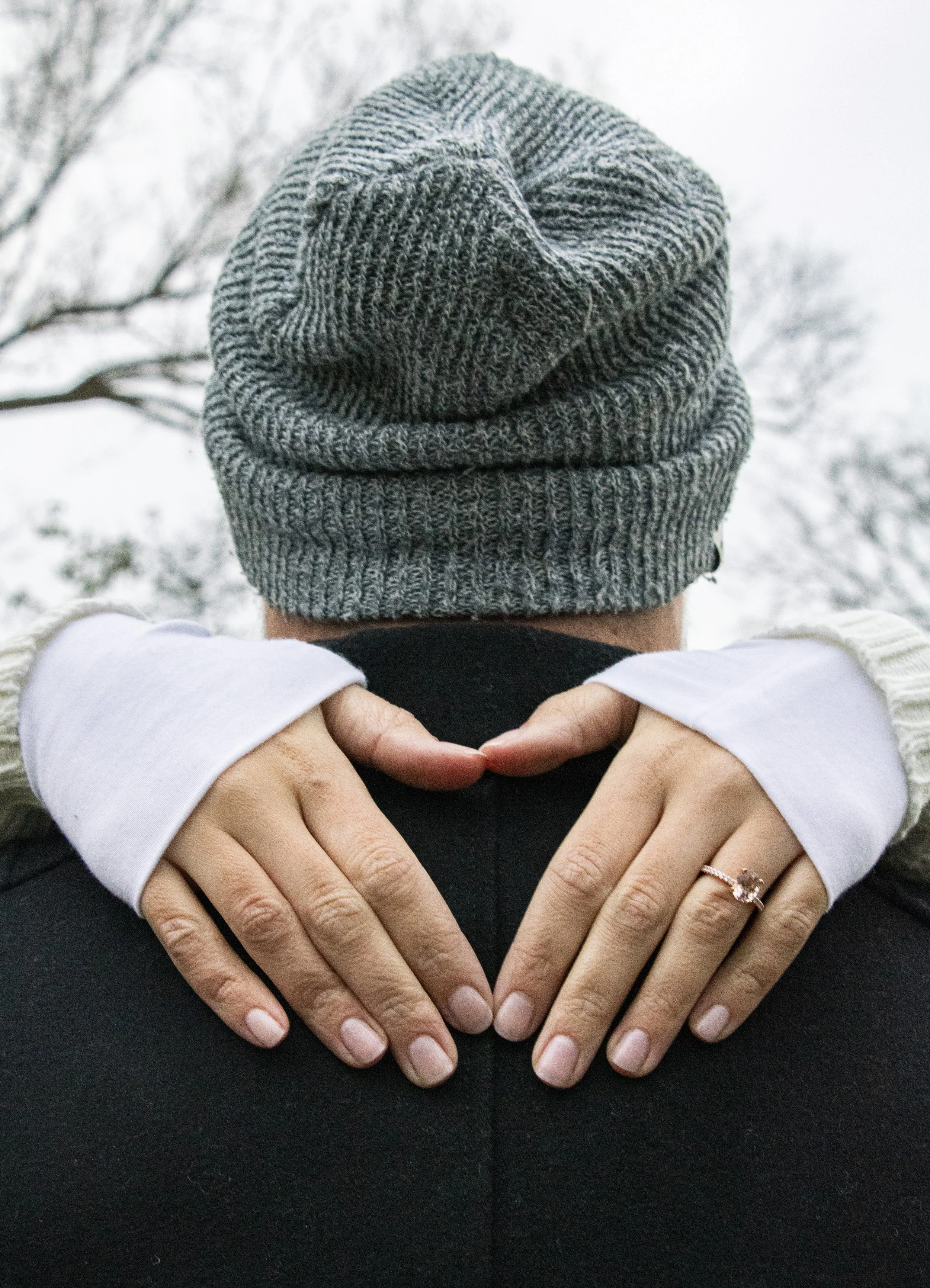 A person with a gray knit beanie and a white long sleeve shirt making a heart shape with their hands on another person's shoulder, outdoors with leafless trees in the background.