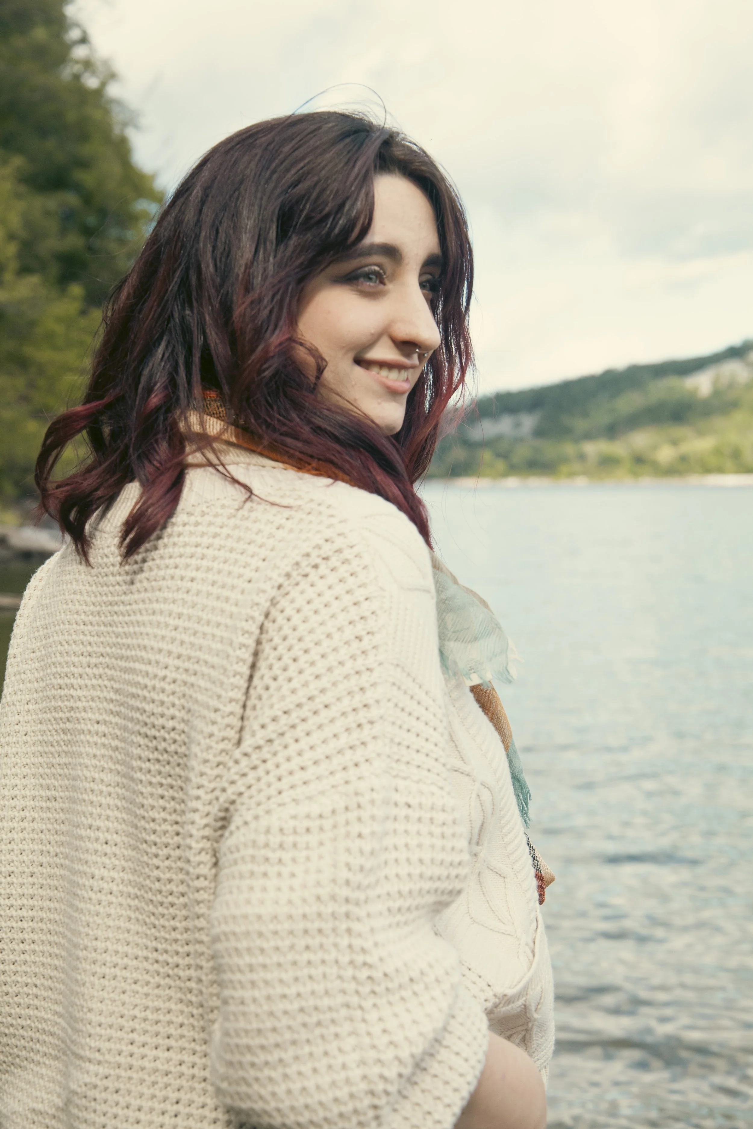 A young woman with dark purple hair and a nose ring smiling near a lake with trees and hills in the background.