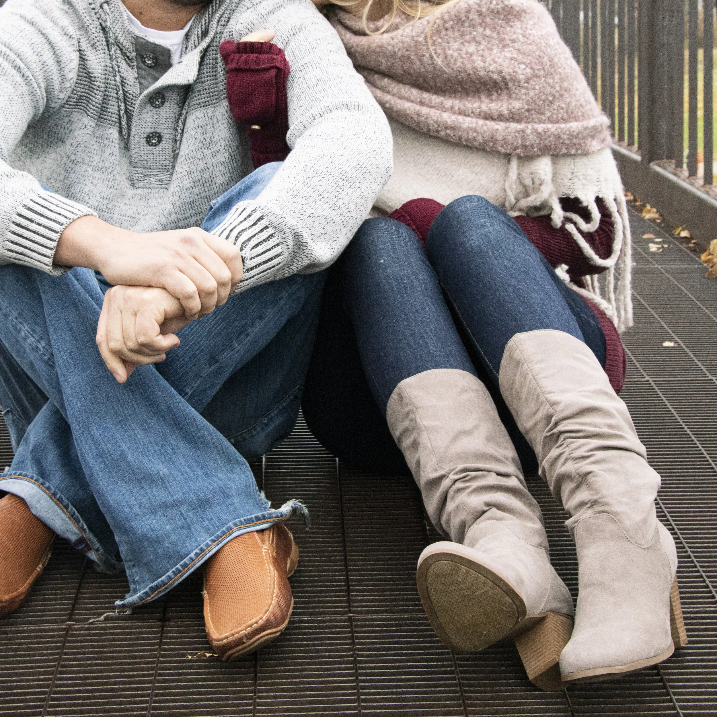 Close-up of two people sitting next to each other on a metal grate, showing their legs and hands, with fallen leaves nearby.