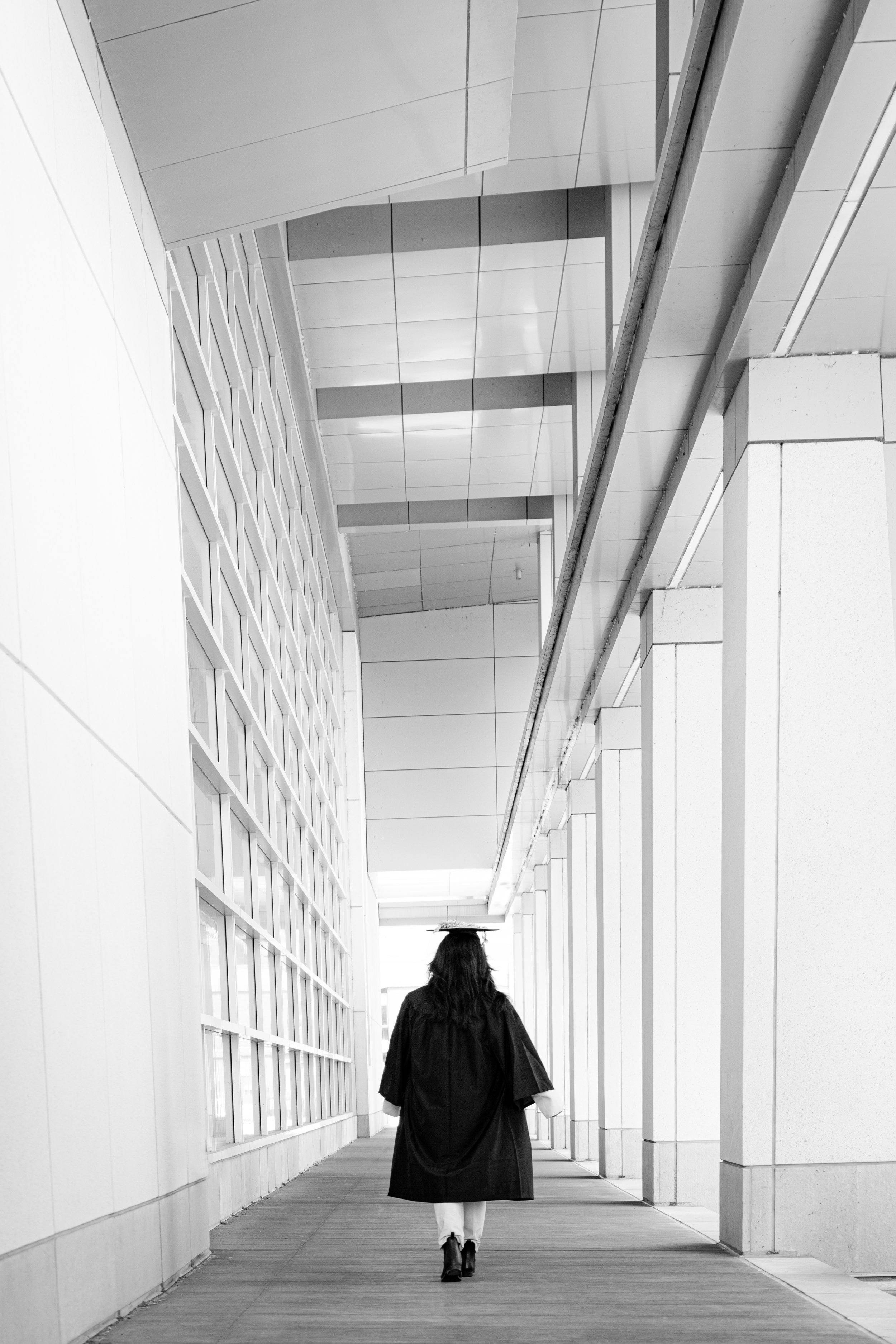 A Hmong person in a graduation gown and cap walking through a modern building corridor with glass walls and structural columns.