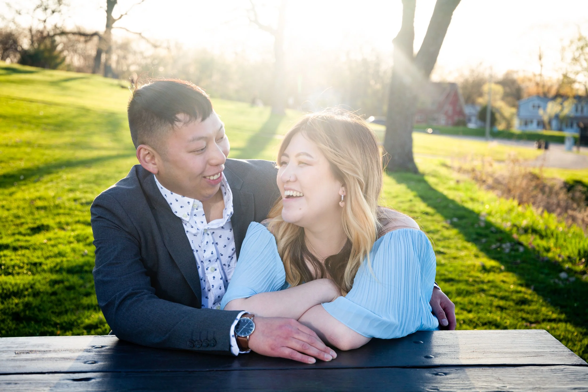 A couple sitting at a wooden table outdoors, smiling and laughing. The man is wearing a dark blazer and patterned shirt, and the woman has long blonde hair and is wearing a light blue blouse. The setting is a park with green grass, trees, and houses 