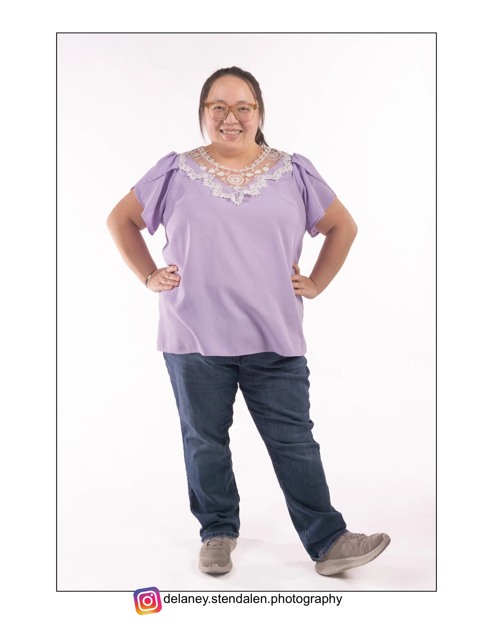 A Hmong woman with glasses and long hair, wearing a lavender blouse with lace details, dark jeans, and sneakers, standing with hands on hips against a white background.