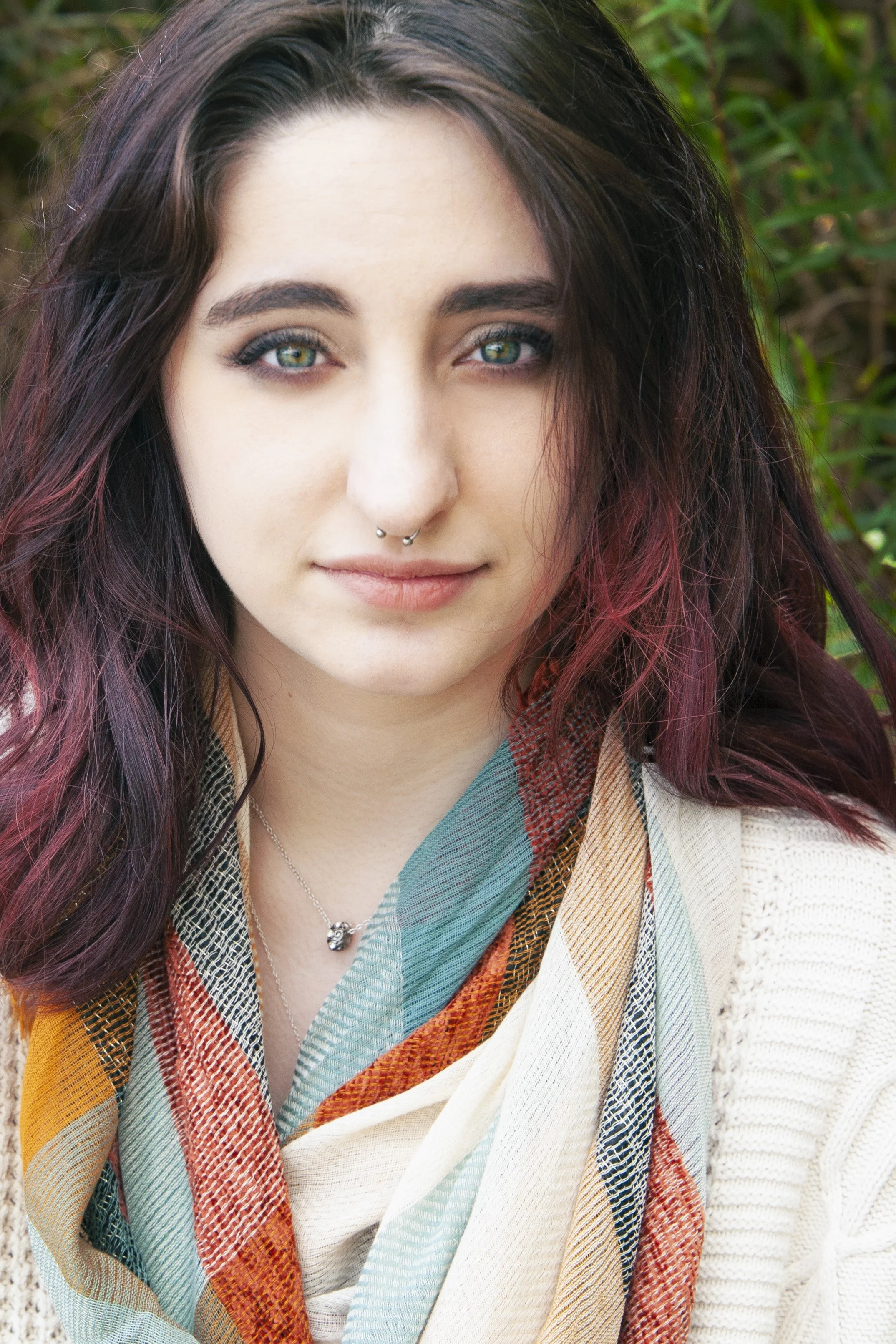 A young woman with long, dark hair with red highlights wearing a colorful scarf and a beige sweater, standing outdoors with greenery in the background.