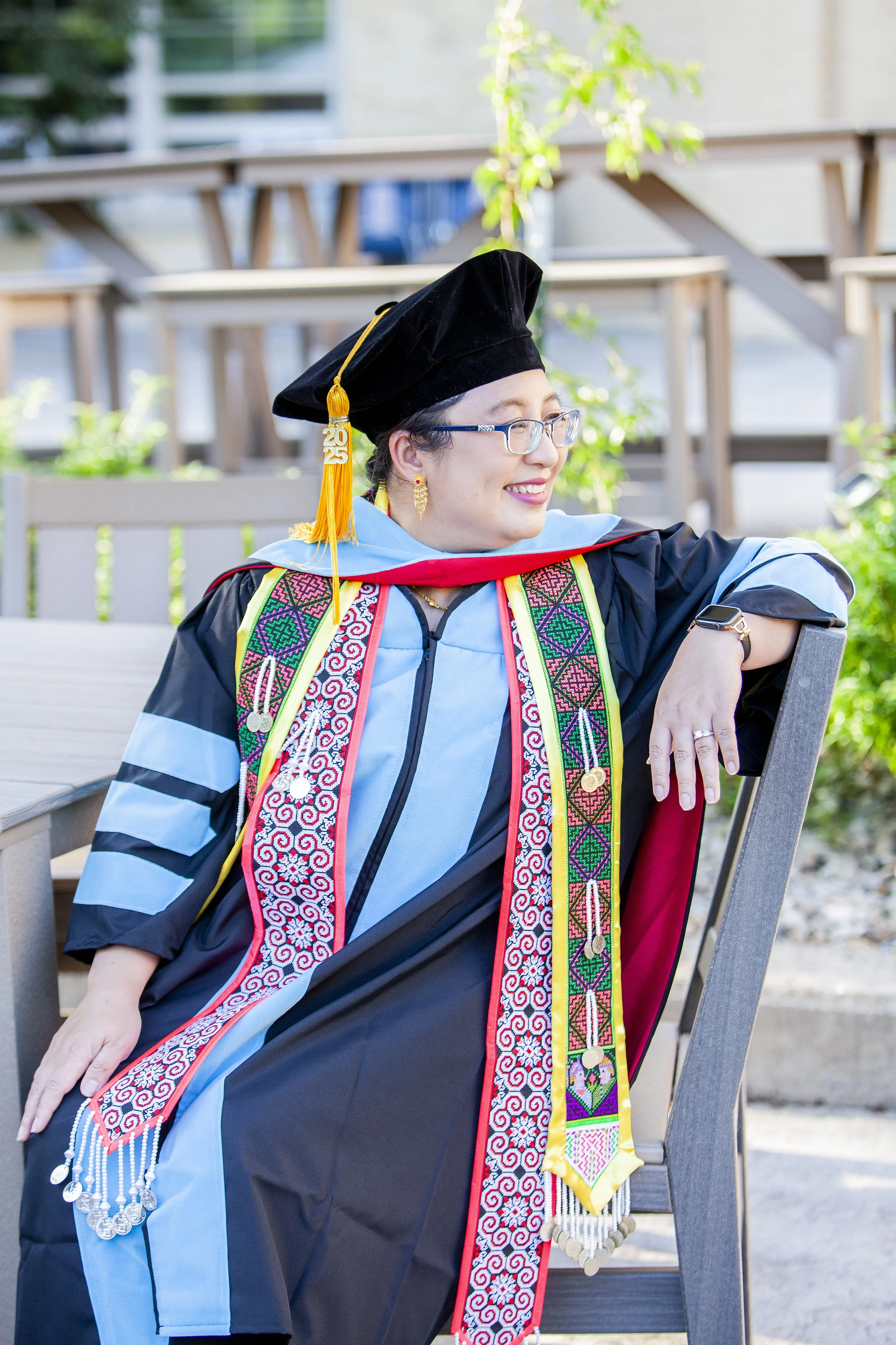 Hmong woman celebrating graduation in academic cap and gown, seated outdoors with greenery and wooden furniture in background.