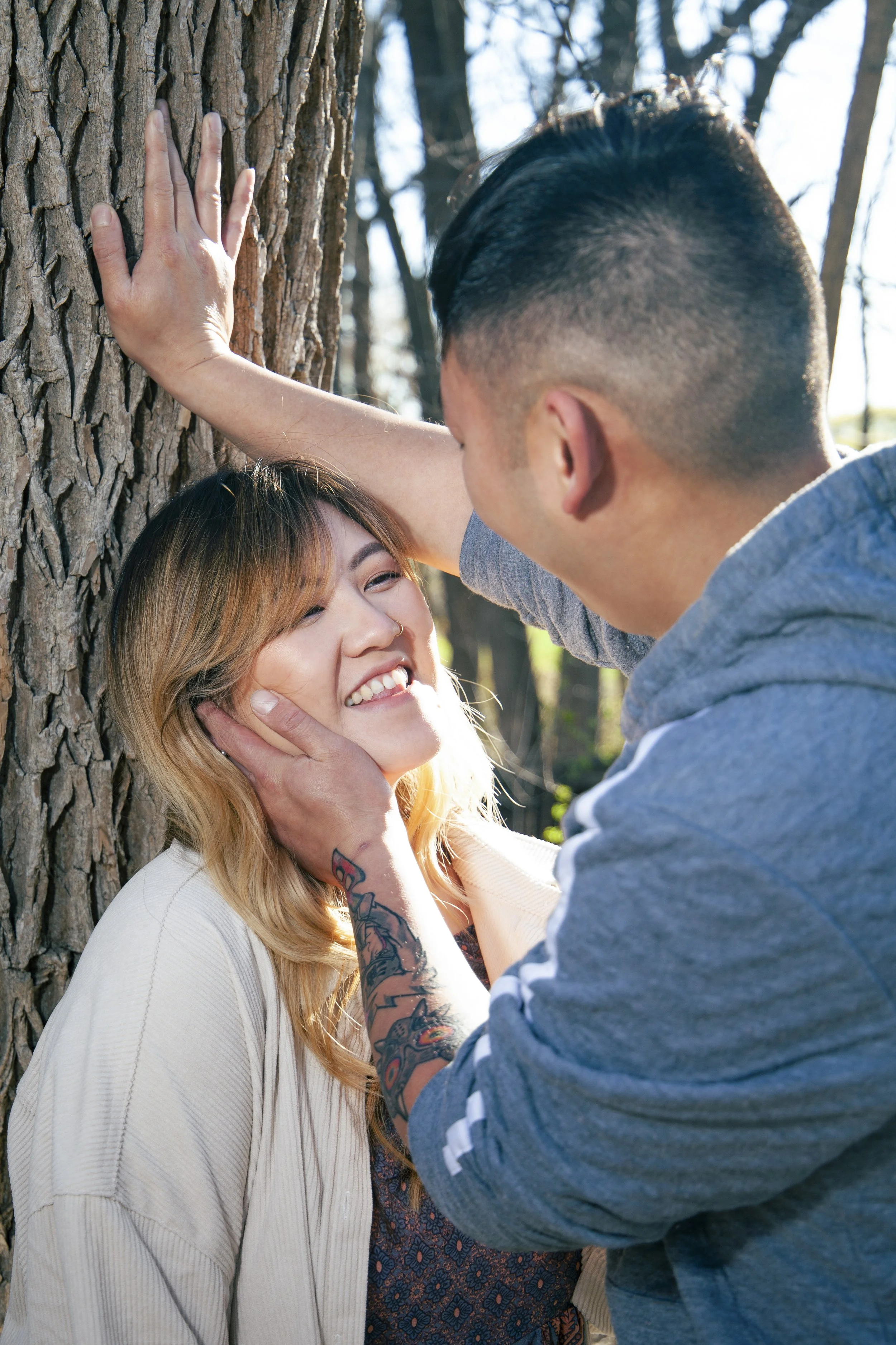A Hmong man and woman are close to each other outdoors, with the man gently holding the woman's face and smiling at her. The woman is smiling, leaning against a tree, and looking happy.