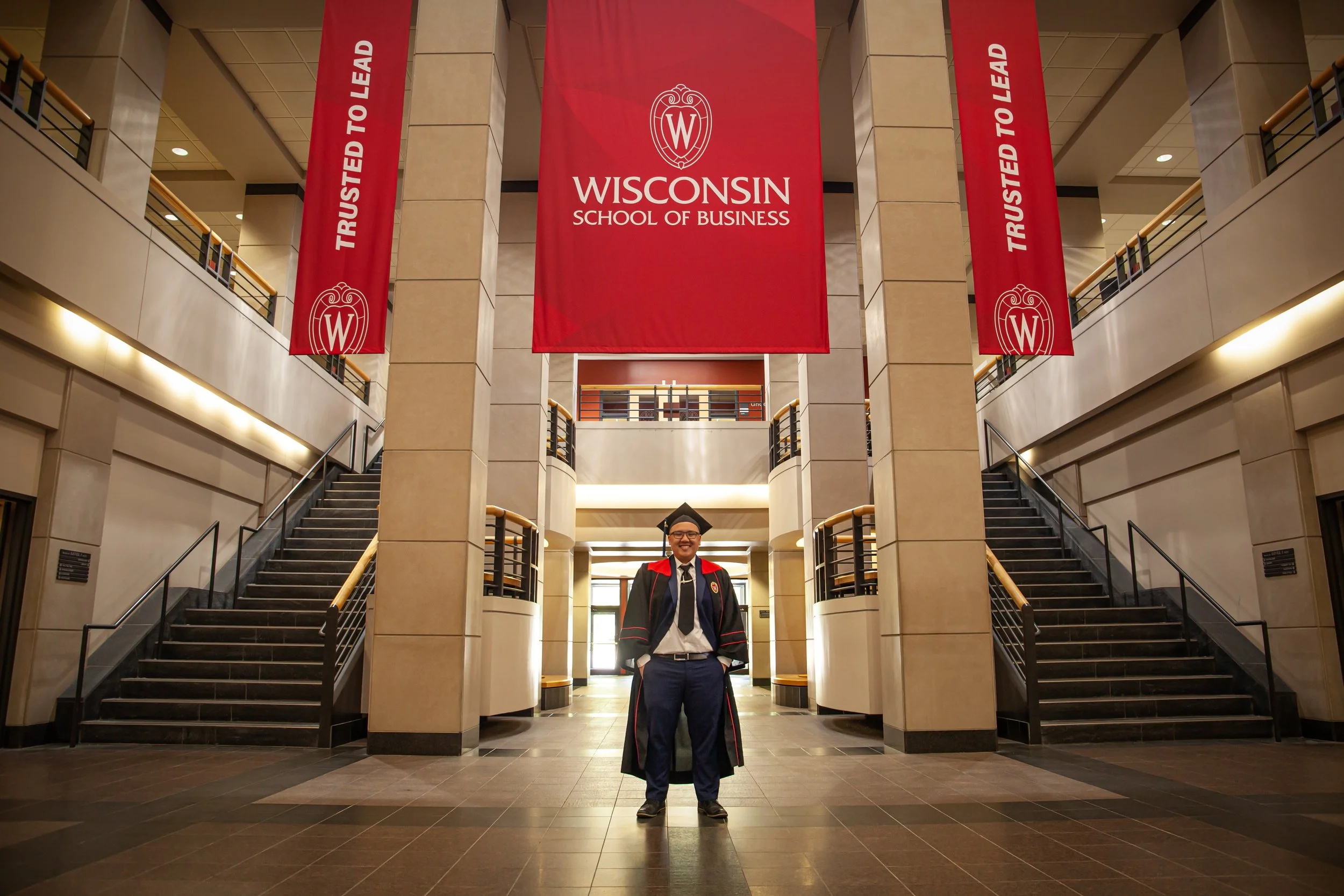 A Hmong graduate in a cap and gown standing inside the Wisconsin School of Business building with banners that say 'Trusted to Lead' and a large red banner displaying the school's name and logo.