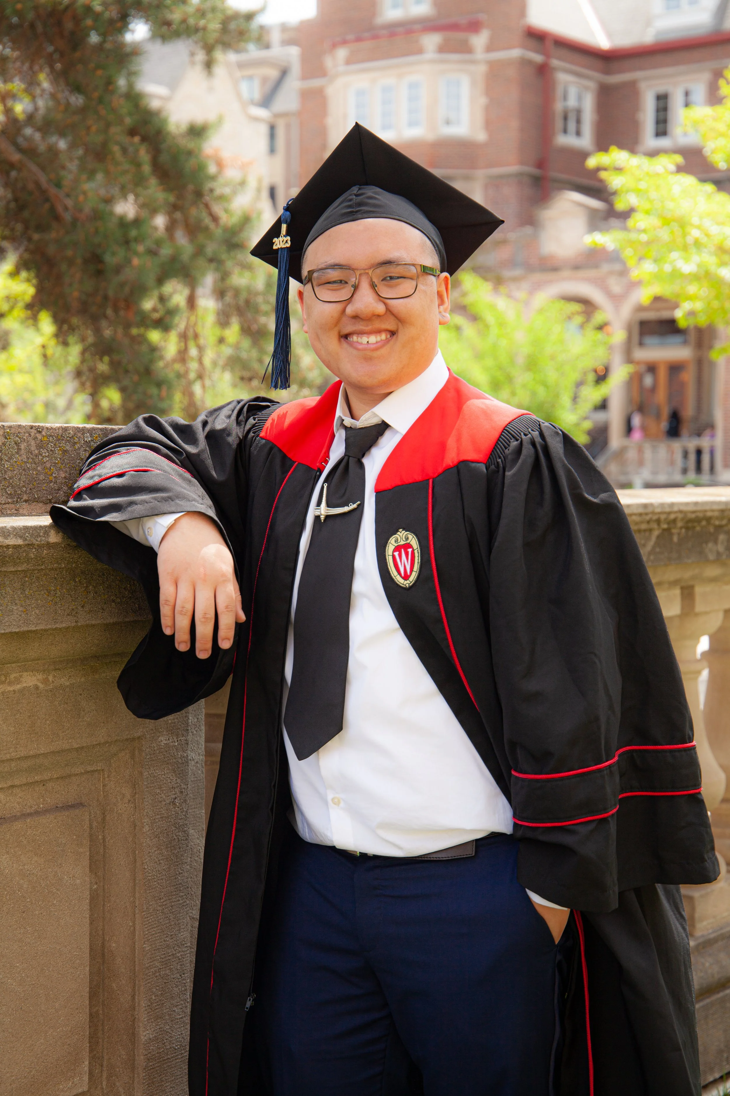A smiling man in graduation cap and gown standing outdoors, leaning on a stone railing with one arm. He is wearing glasses, a white shirt, black tie, and has a WI logo badge on his gown. The background features trees and a historic brick building.