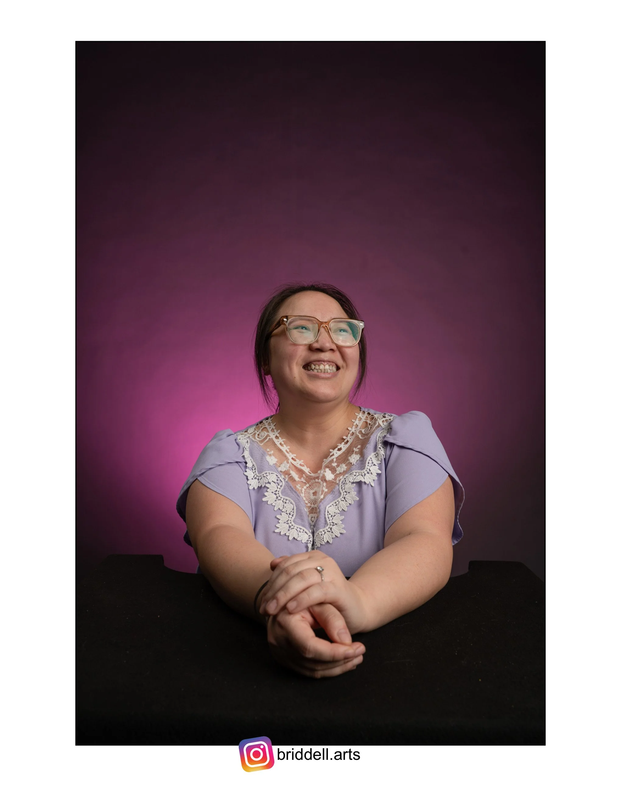 Hmong woman smiling, wearing glasses and a lavender blouse with lace detail, sitting at a table against a pink and purple gradient background.
