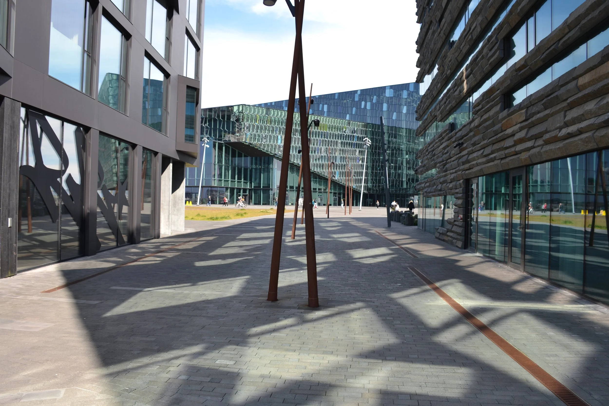 Pedestrian approach to Harpa Concert Hall with sunlight and shadows patterning the walkway between reflective buildings.